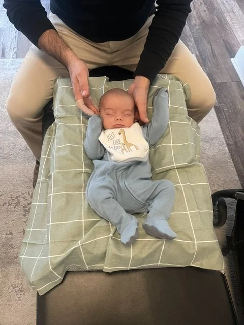 A person holding a sleeping baby on a changing table, with the baby's arms raised and wearing a bib and blue pants, in an indoor setting.