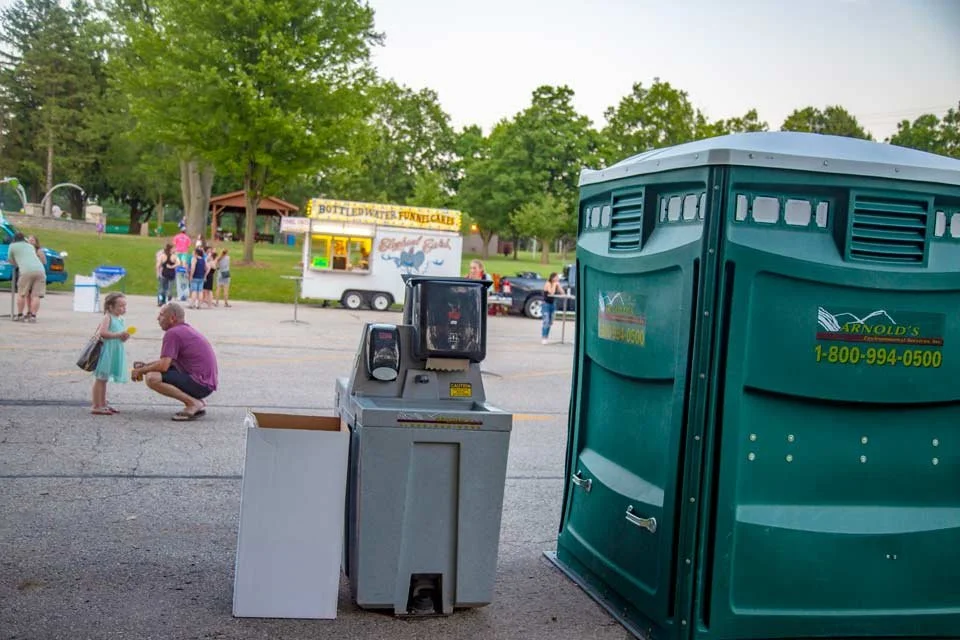 A small girl and an older man in purple shorts and a purple shirt shaking hands in an outdoor setting near a food stand, with portable toilets in the foreground, and people in the background at a park.