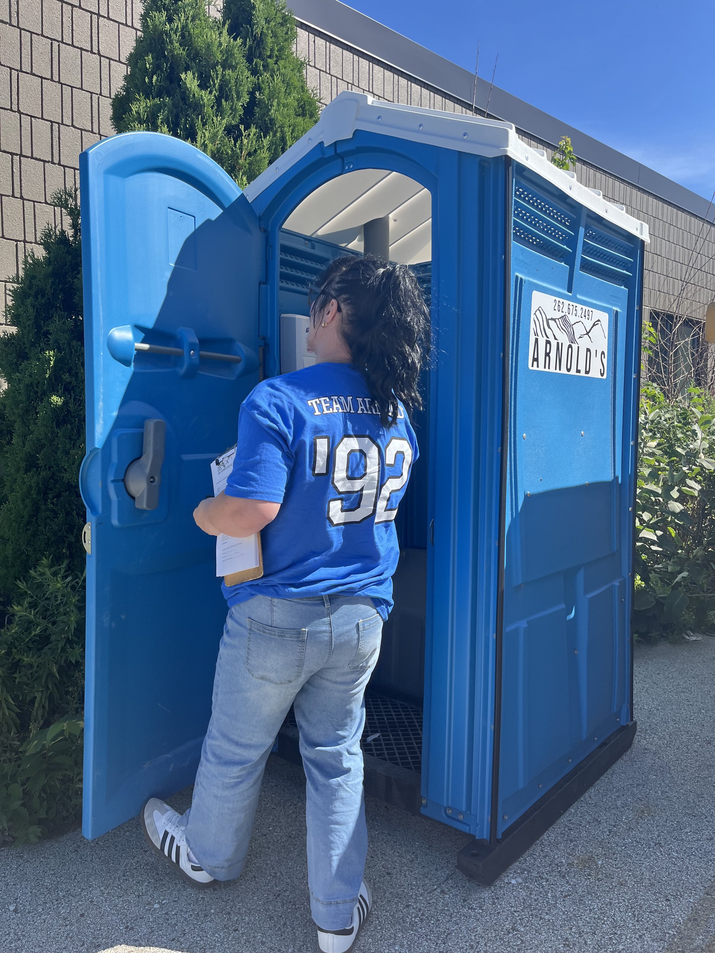 A person with dark hair wearing a blue sports jersey and light jeans is kneeling in front of a blue portable restroom, looking inside with the door slightly open under a clear blue sky.