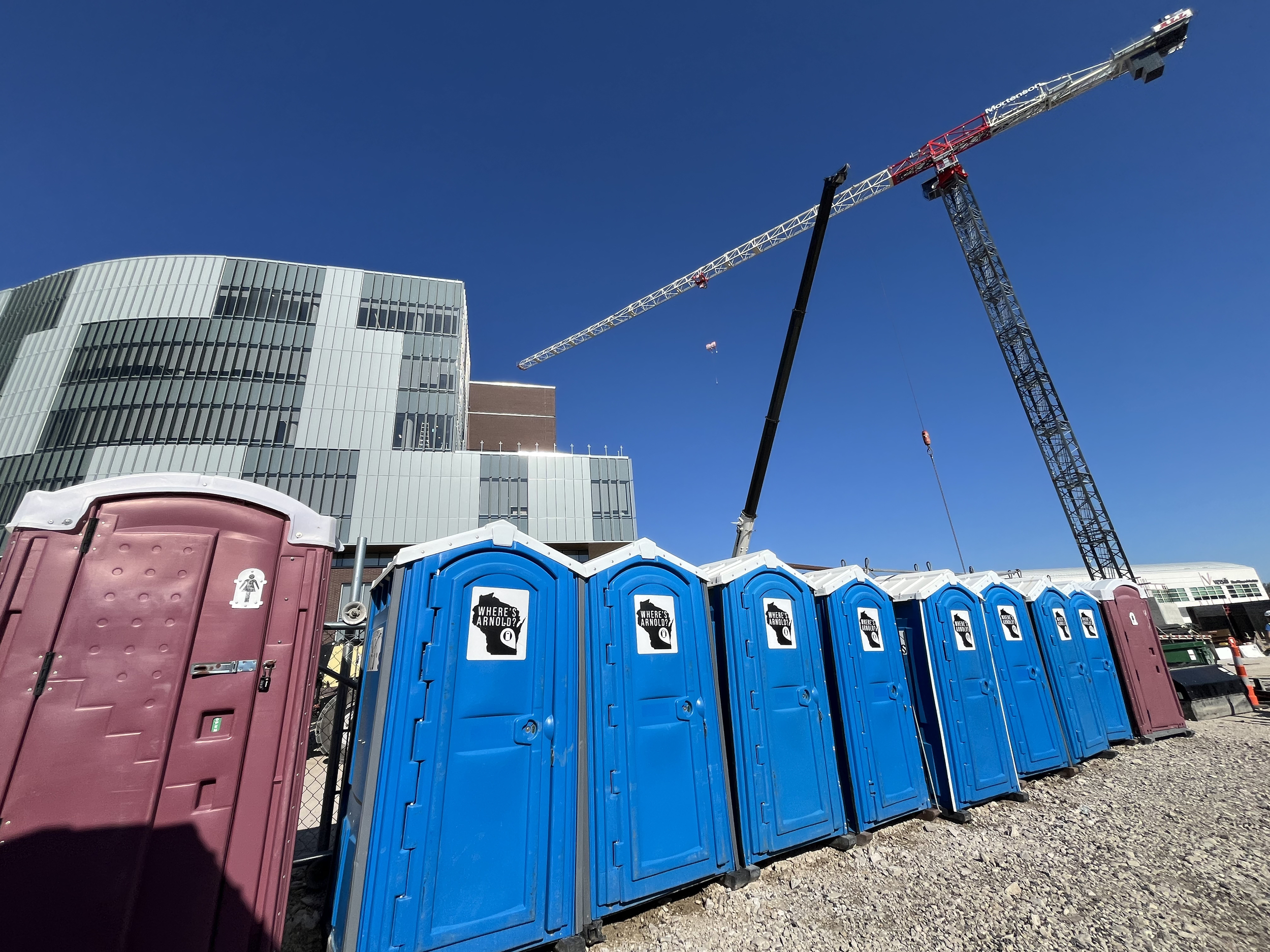 A bank of Arnold's portable restrooms on a Wisconsin construction site, including blue units and a pink women's unit, with a large crane towering overhead.