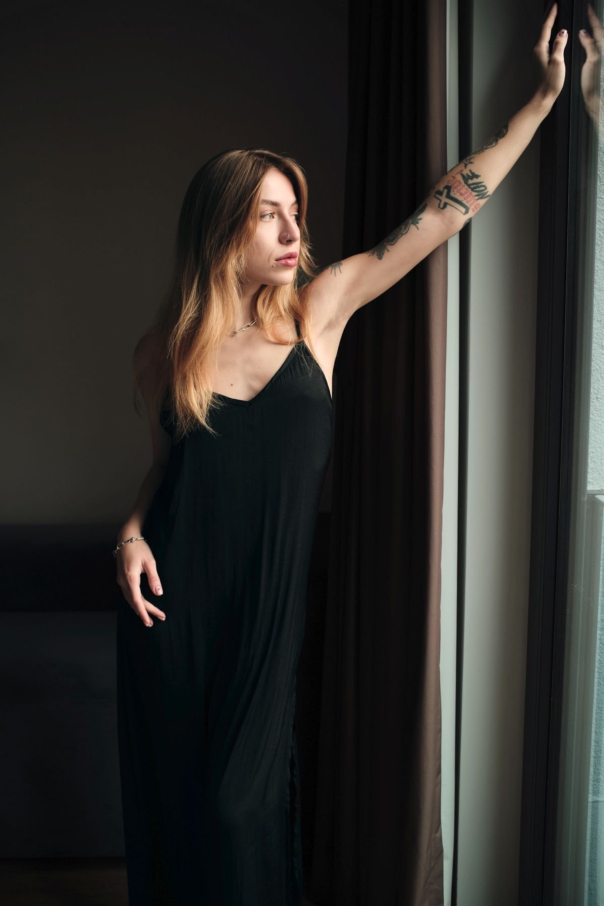 Junge Frau mit Tattoos trägt schwarzes Kleid und steht am Fenster, schaut nach außen, berührt den Vorhang.