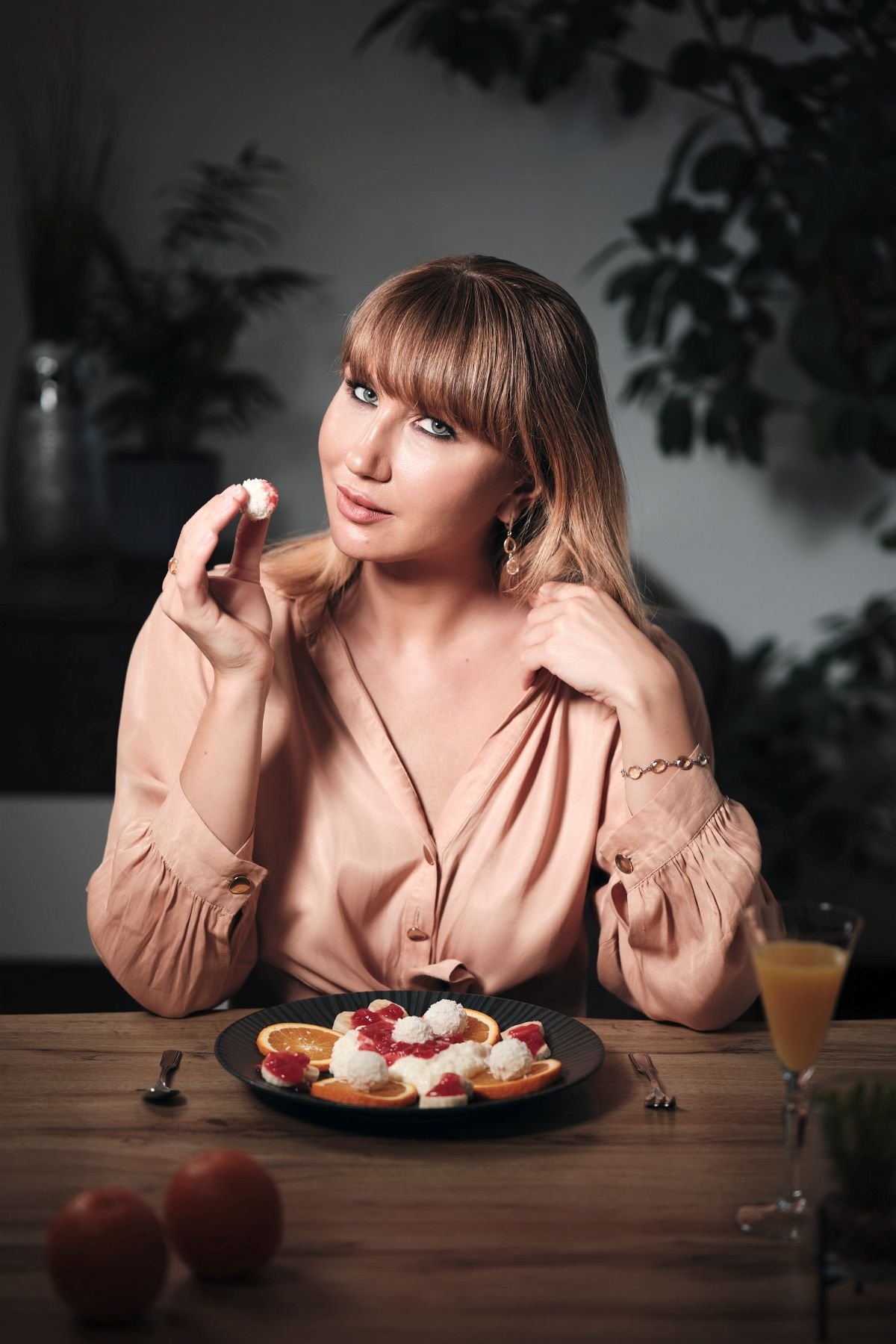 Eine Frau mit braunen Haaren und blauer Augen, die einen Löffel mit Kokosnuss in der Hand hält. Vor ihr auf dem Tisch liegt ein Teller mit einem Dessert aus Orangen, Kirschen und Kokosnüssen. Sie trägt eine beige Seidenbluse und elegante Ohrringe.