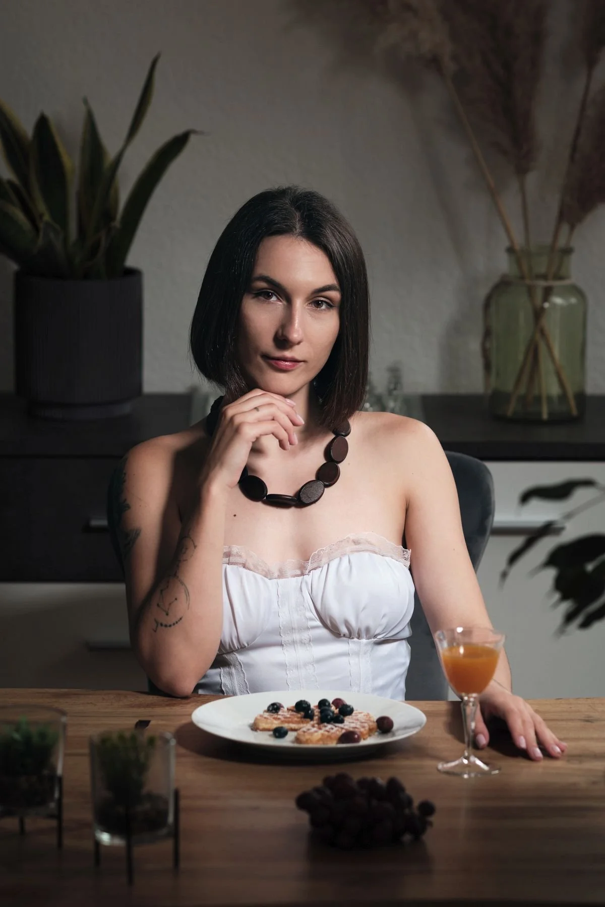 Eine junge Frau mit dunklem Haar und einem schwarzen Halskettchen sitzt an einem Holztisch, vor ihr ein Teller mit Waffeln und Beeren, ein Cocktailglas mit orangefarbener Flüssigkeit, im Hintergrund Topfpflanzen.