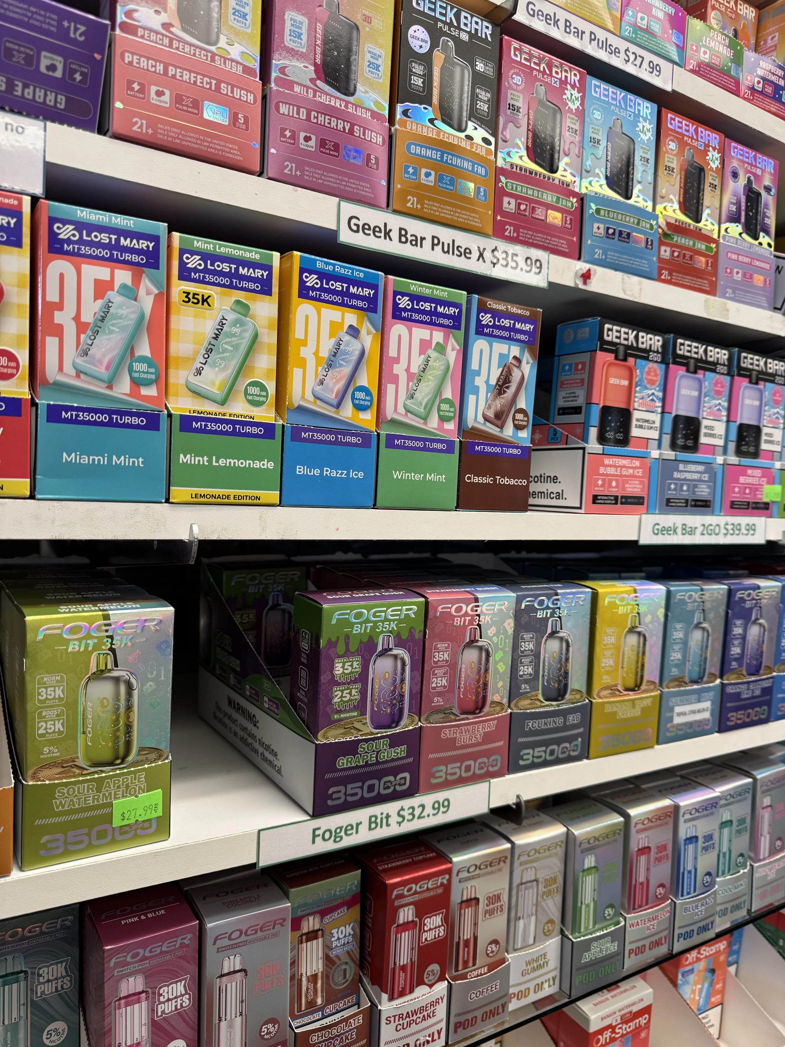 Shelves displaying various vape products with brands like Geek Bar and Lost Mary, showcasing different flavors and nicotine levels. Prominent price tags and warnings about nicotine addiction are visible.