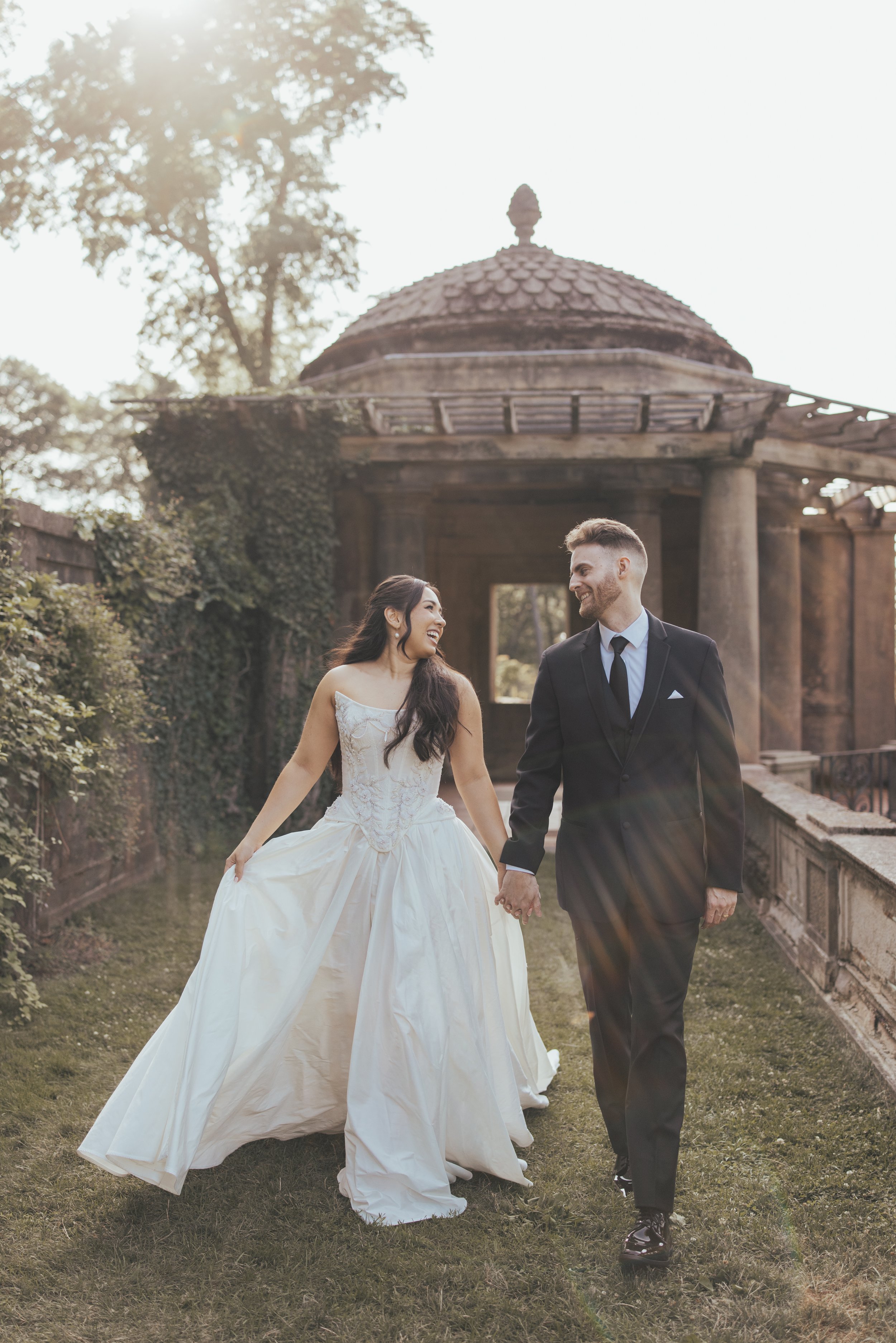A bride and groom smiling happily at their wedding outdoors, with greenery in the background, the bride holding a bouquet of white and lavender flowers.