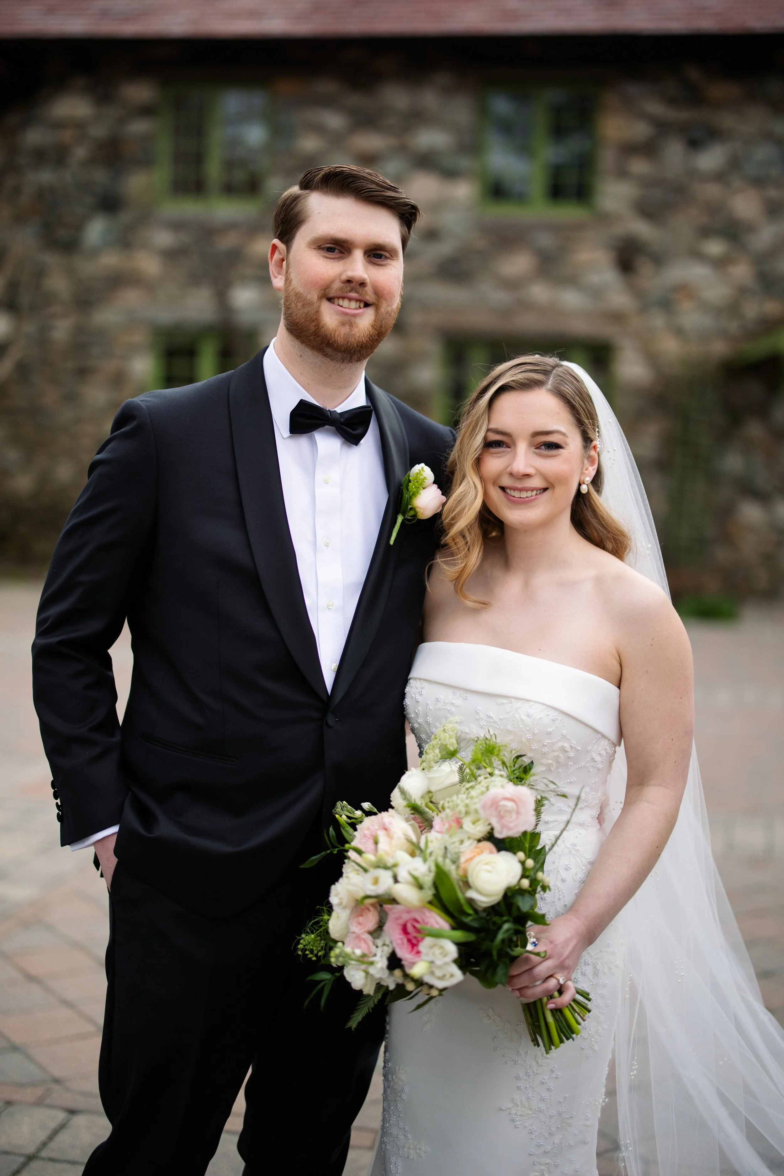 A smiling bride and groom standing together outdoors on their wedding day. The groom is in a black tuxedo with a bow tie, and the bride is in a strapless white wedding gown holding a bouquet of white and pink flowers. They are in front of a stone building with green window frames.