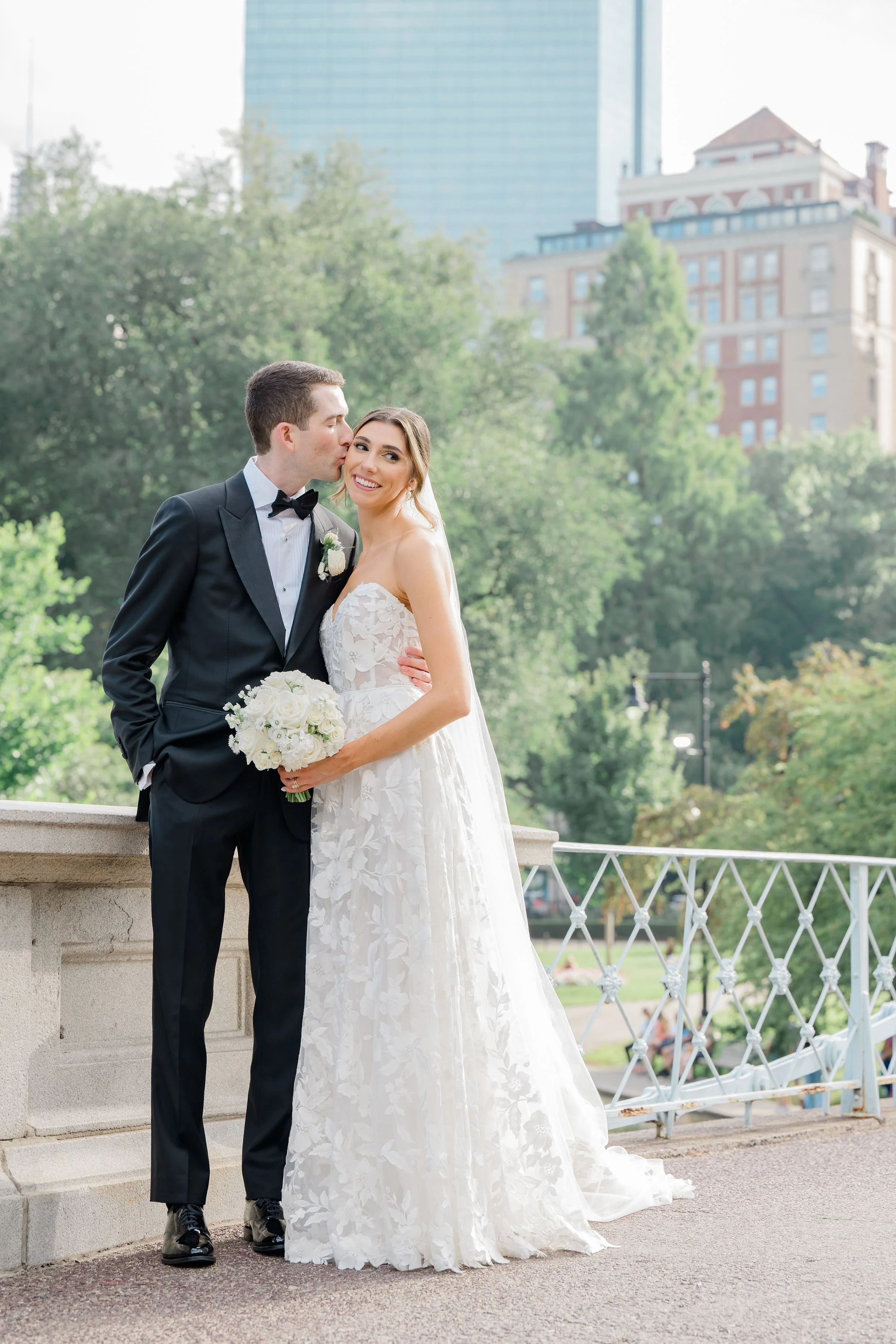 A newlywed couple outdoors in a park, with the groom in a black tuxedo and the bride in a white lace wedding dress holding a bouquet of white flowers. The groom is kissing the bride on her temple, and she is smiling. There are trees and tall buildings in the background.