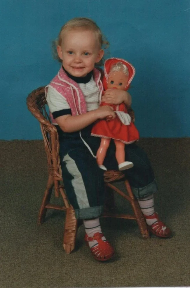 Young child sitting on a wicker chair holding a vintage doll with blonde hair, wearing a pink sweater vest, striped shirt, jeans with rolled-up cuffs, striped socks, and red sandals, against a blue background.