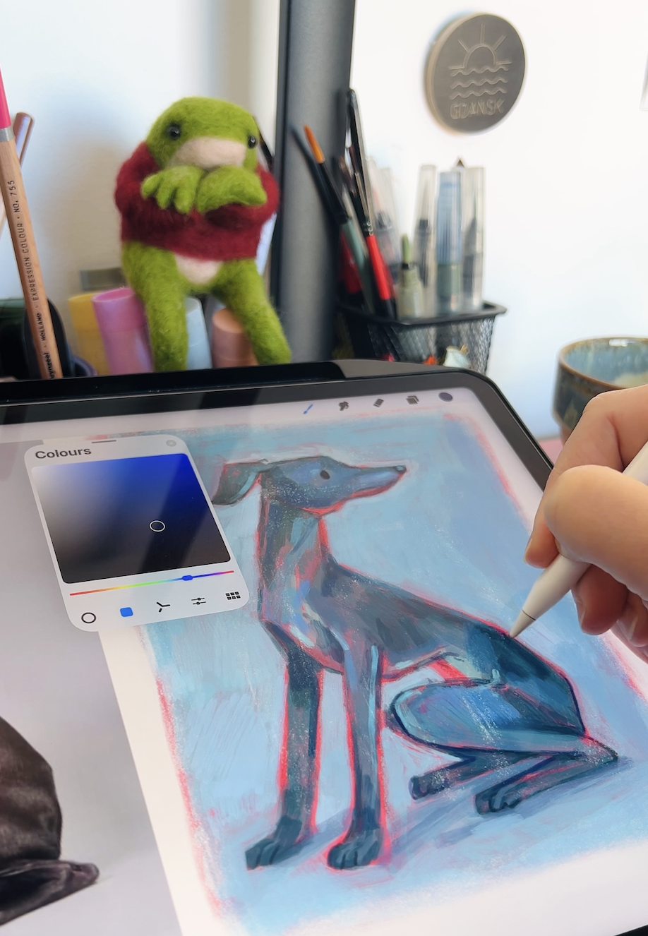 Digital drawing of a sitting dog with a stylized look on a tablet screen, with an artist's hand using a stylus to add details, in a workspace with art supplies and a plush frog toy in the background.