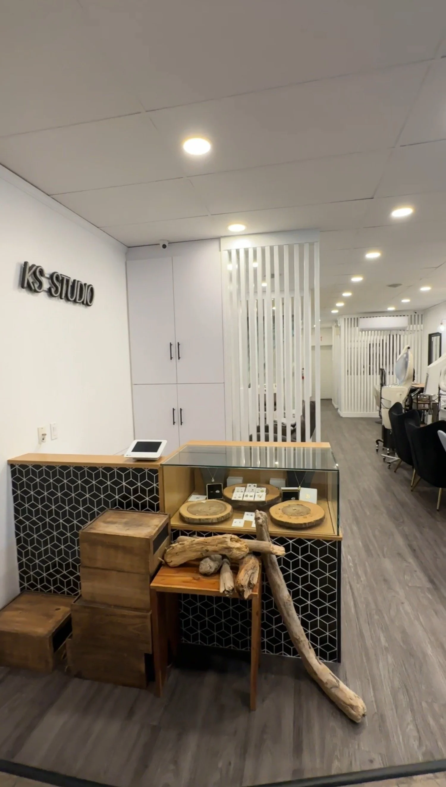 Interior of a modern salon or studio with a display case containing jewelry, wooden decorative pieces, and natural wood logs and branches in front of it. The space has white walls, cabinetry, black and white geometric decor, and black chairs.
