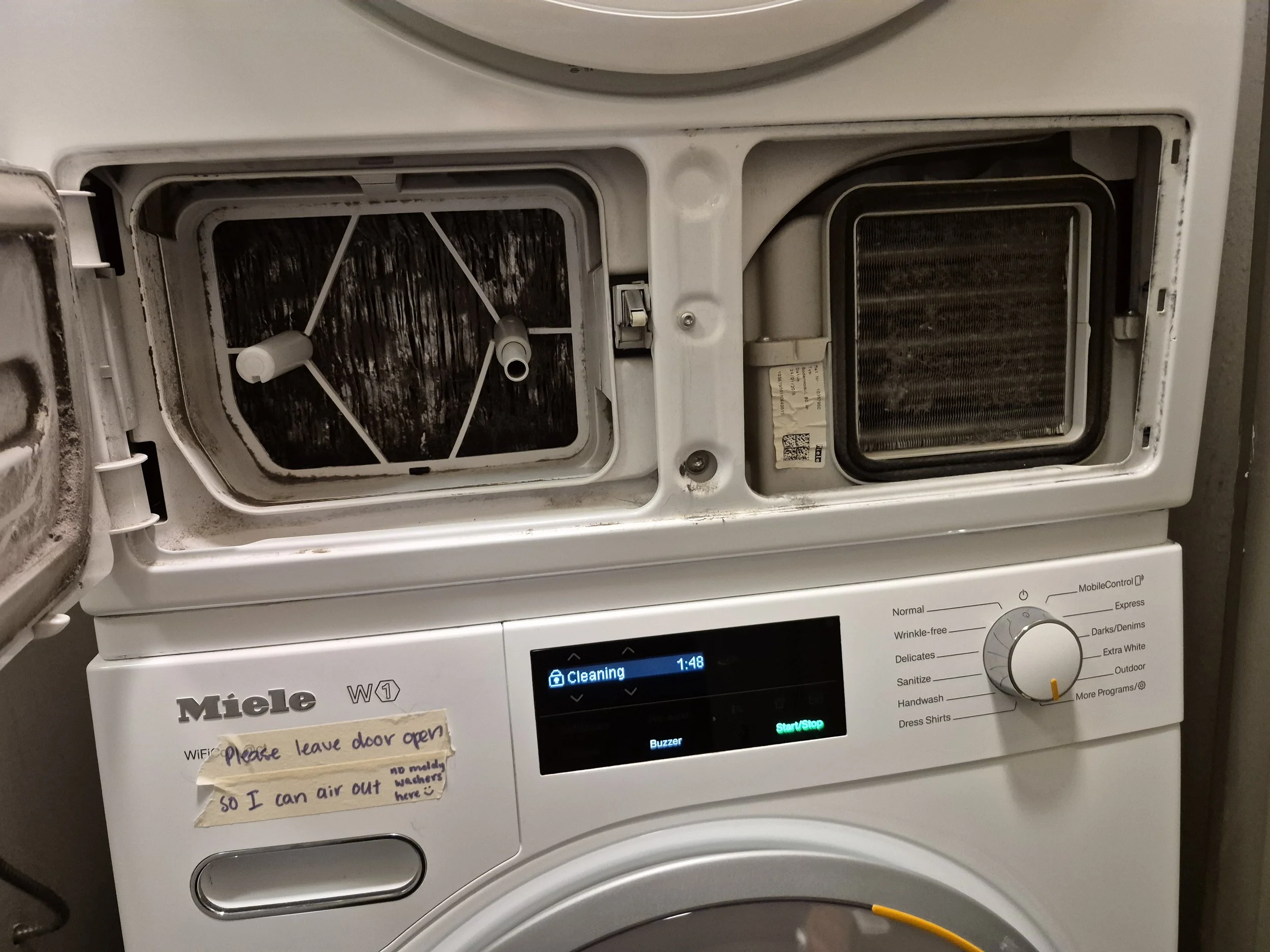 An open front-loading washing machine with the door left ajar. The washing machine has a digital display showing the time as 1:48 and various settings. There is a handwritten note taped to the front of the machine that reads: 'Please leave door open 