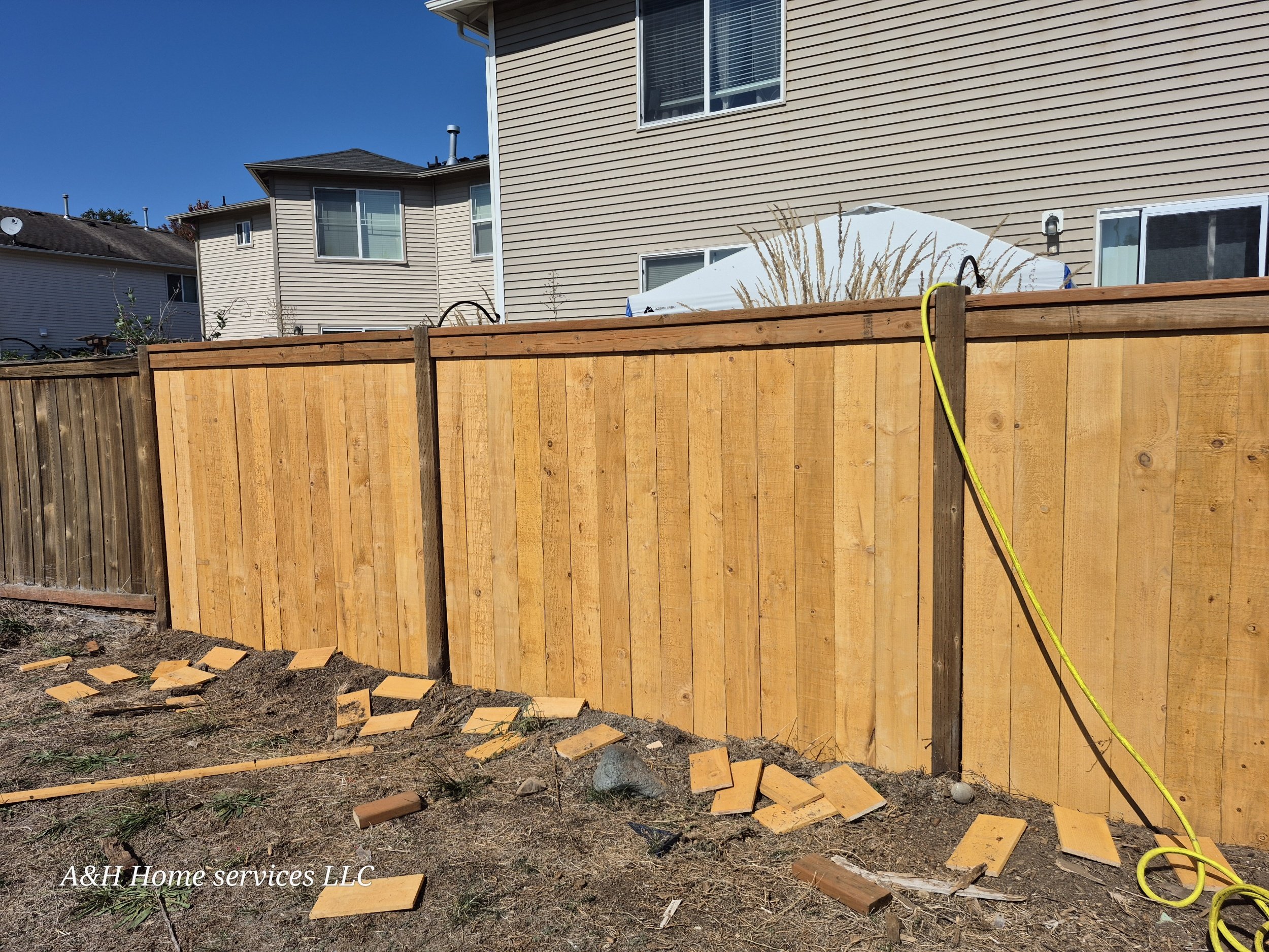 New wooden fence under construction in a backyard, with some loose fence panels and tools on the ground, adjacent to existing fences and houses.