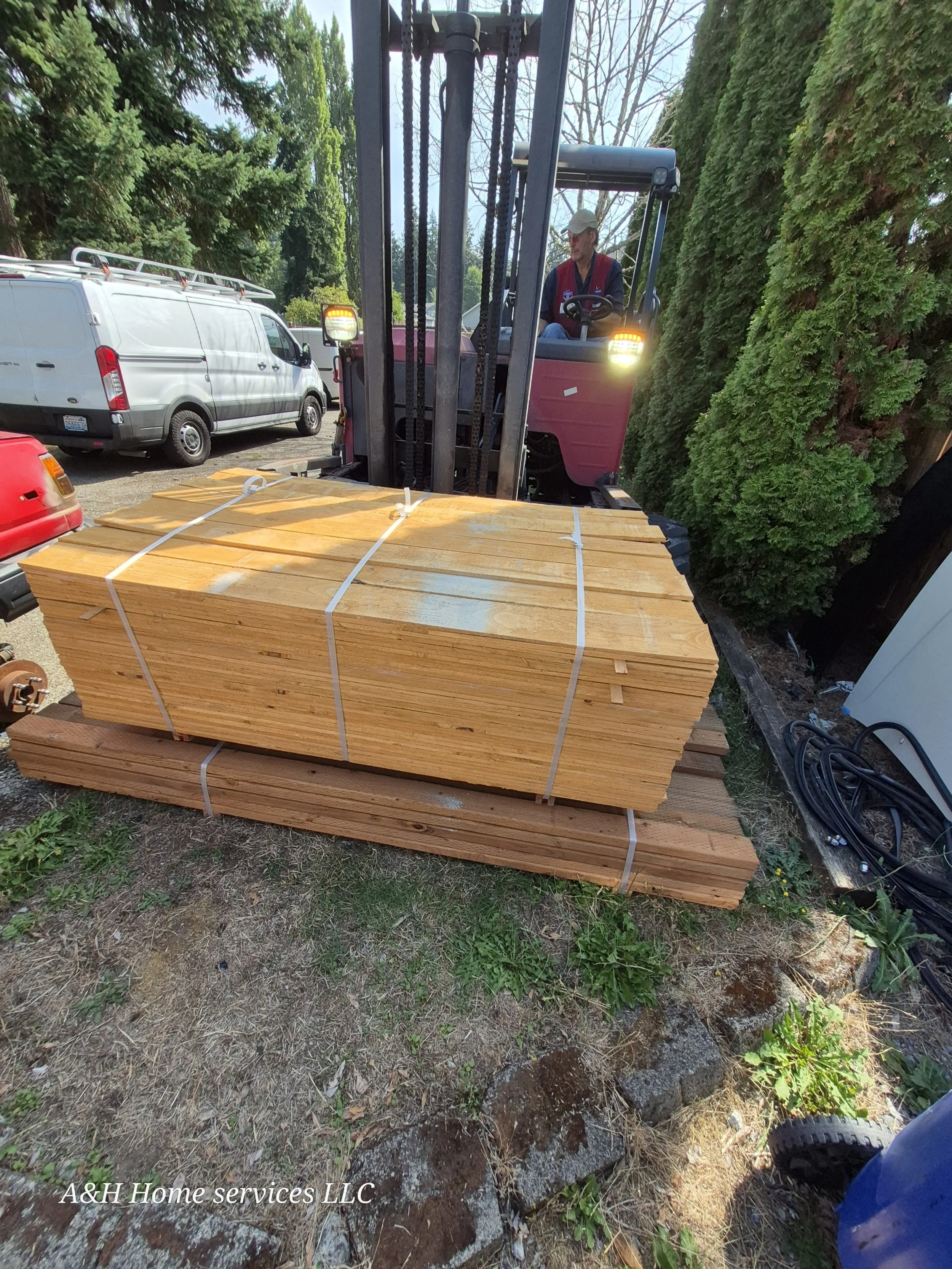 Pallet of cut lumber on the ground with a forklift and a person operating it, outdoor setting with trees and vehicles nearby.