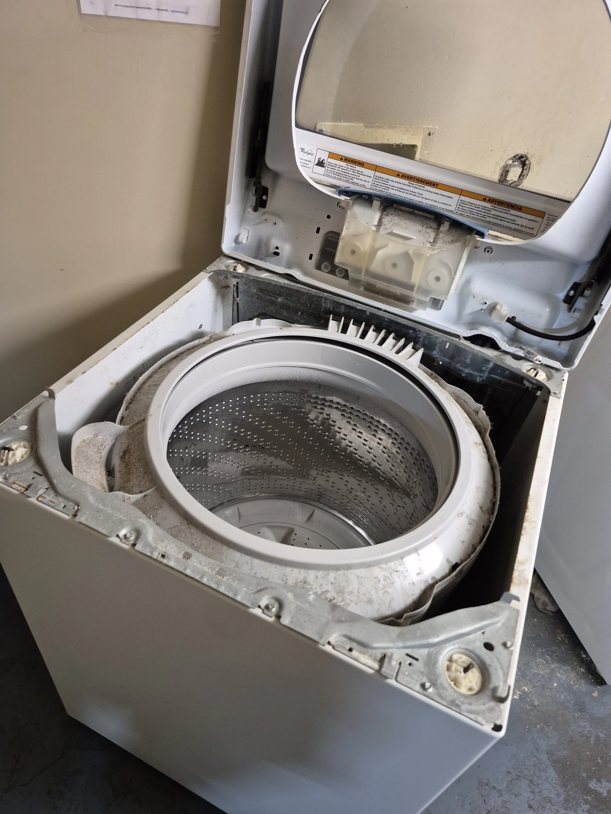 Open top-loading washing machine with visible drum and detached top lid, showing interior and some dirt or dust inside.