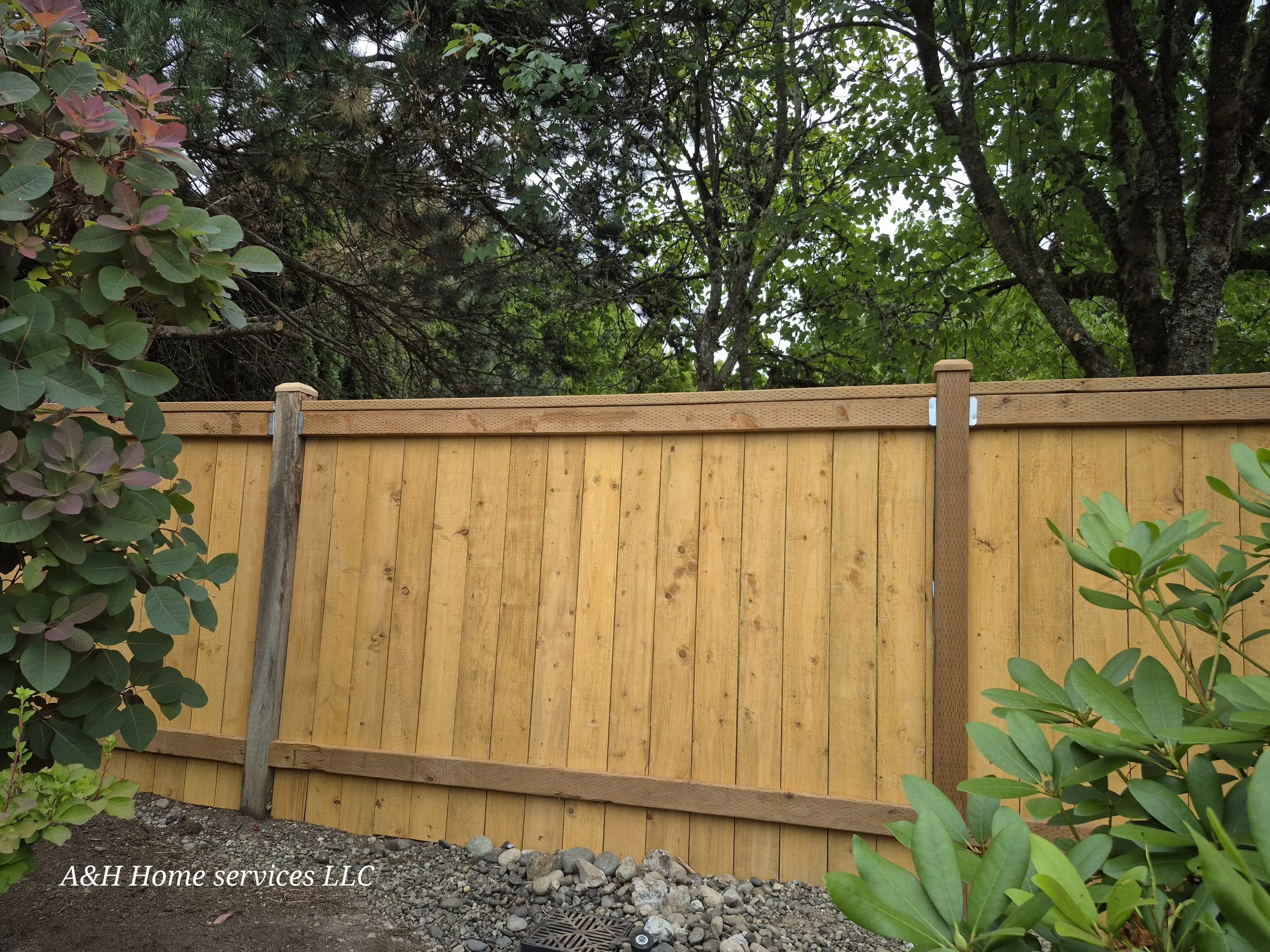Wooden privacy fence with metal brackets, surrounded by green bushes and trees, outdoor setting with visible rocks and soil at the base.