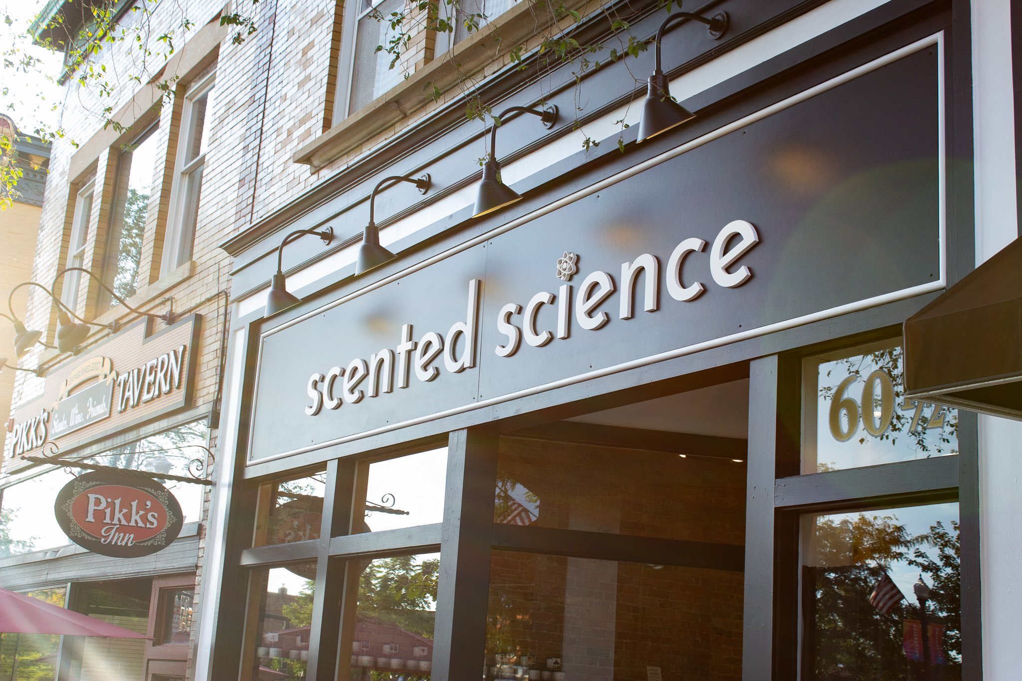  storefront with signs for scented science and Pikk's Inn, along a brick building with black trim, and trees reflected in the glass