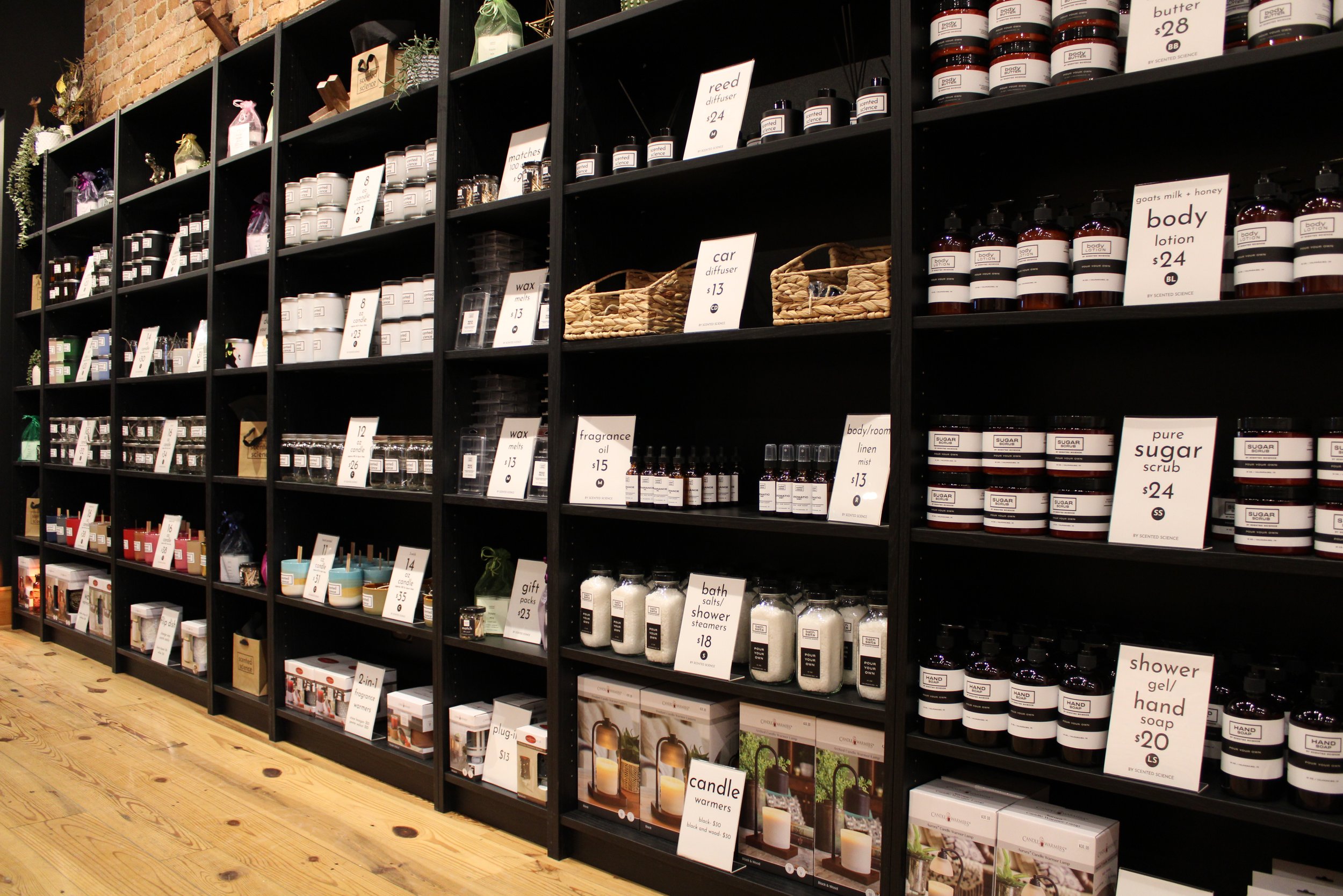 Black shelving unit with various candles, diffusers, lotions, and bath products arranged in rows, with price tags and signs indicating different scents and products.