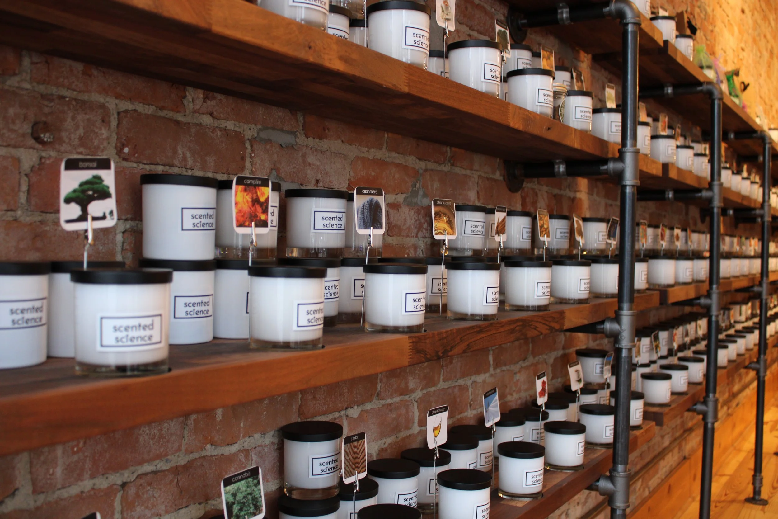 Shelf display of scented candles in white containers with scent labels and small tags showing various scents like cedar, champagne, and cannabis, against a brick wall.
