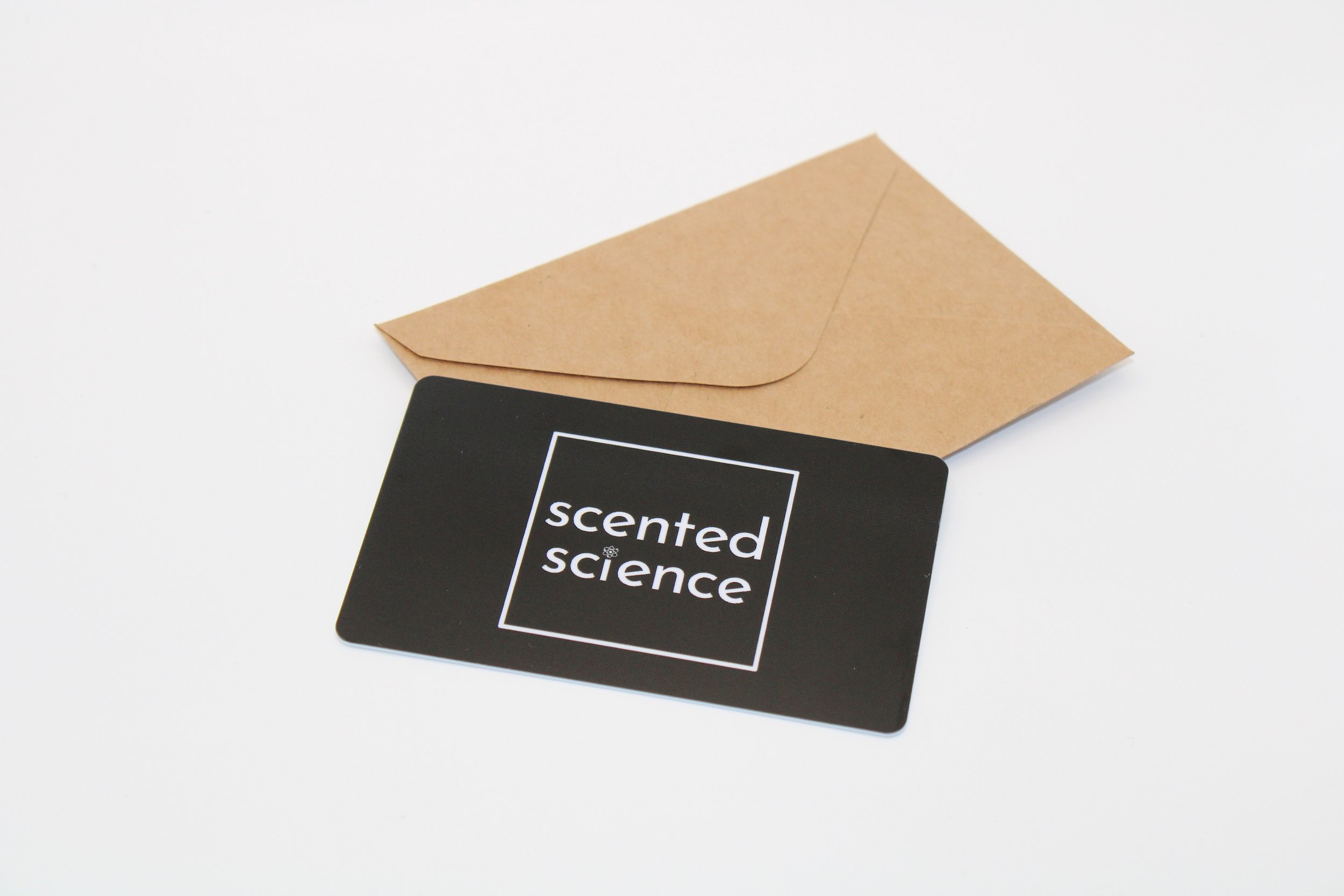 A black card with the words "scented science" inside a white square, and a plain brown envelope behind it, placed on a white surface.