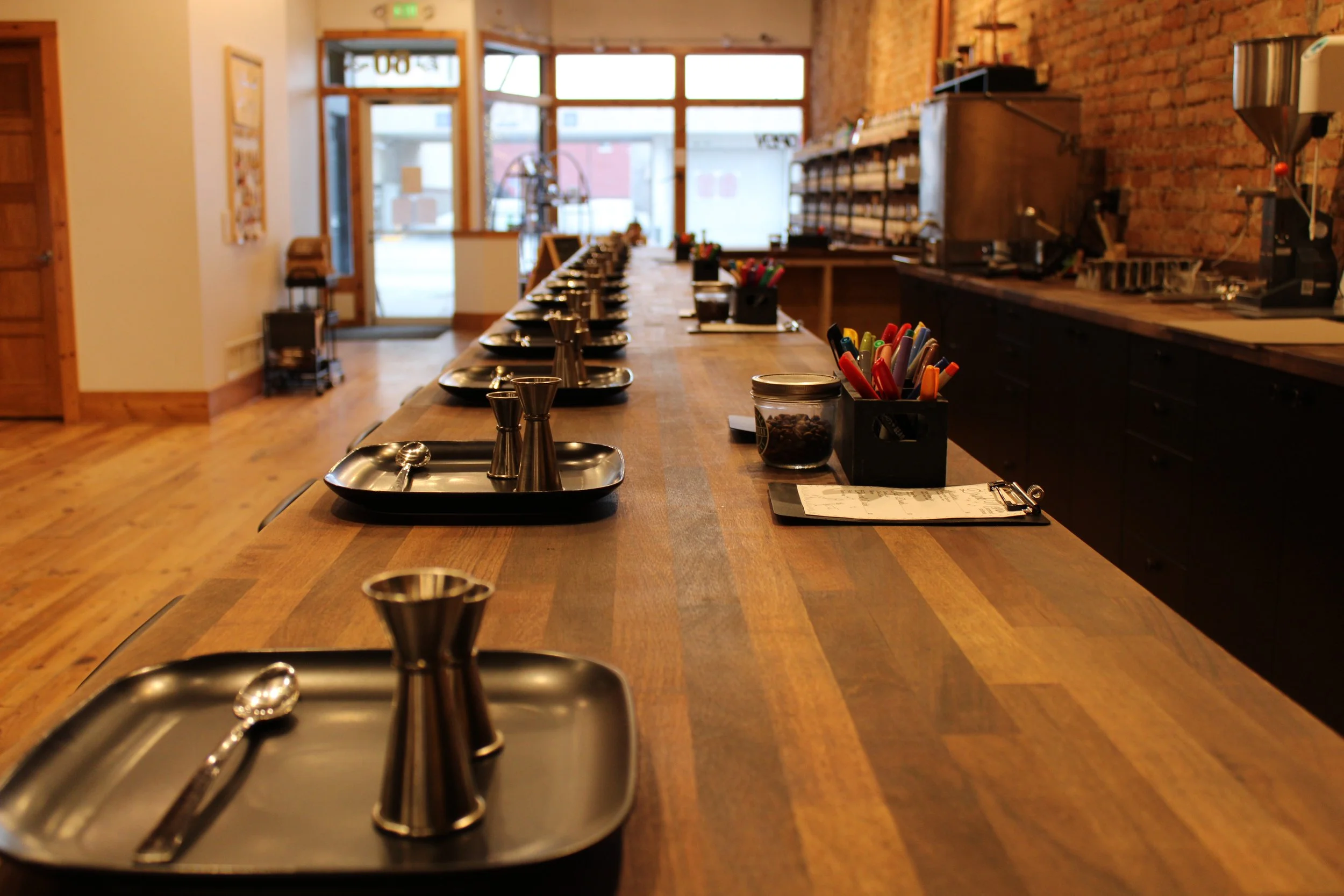 Empty cafe or restaurant counter with trays, jiggers, and utensils set for a coffee or drink service, with large windows in the background.