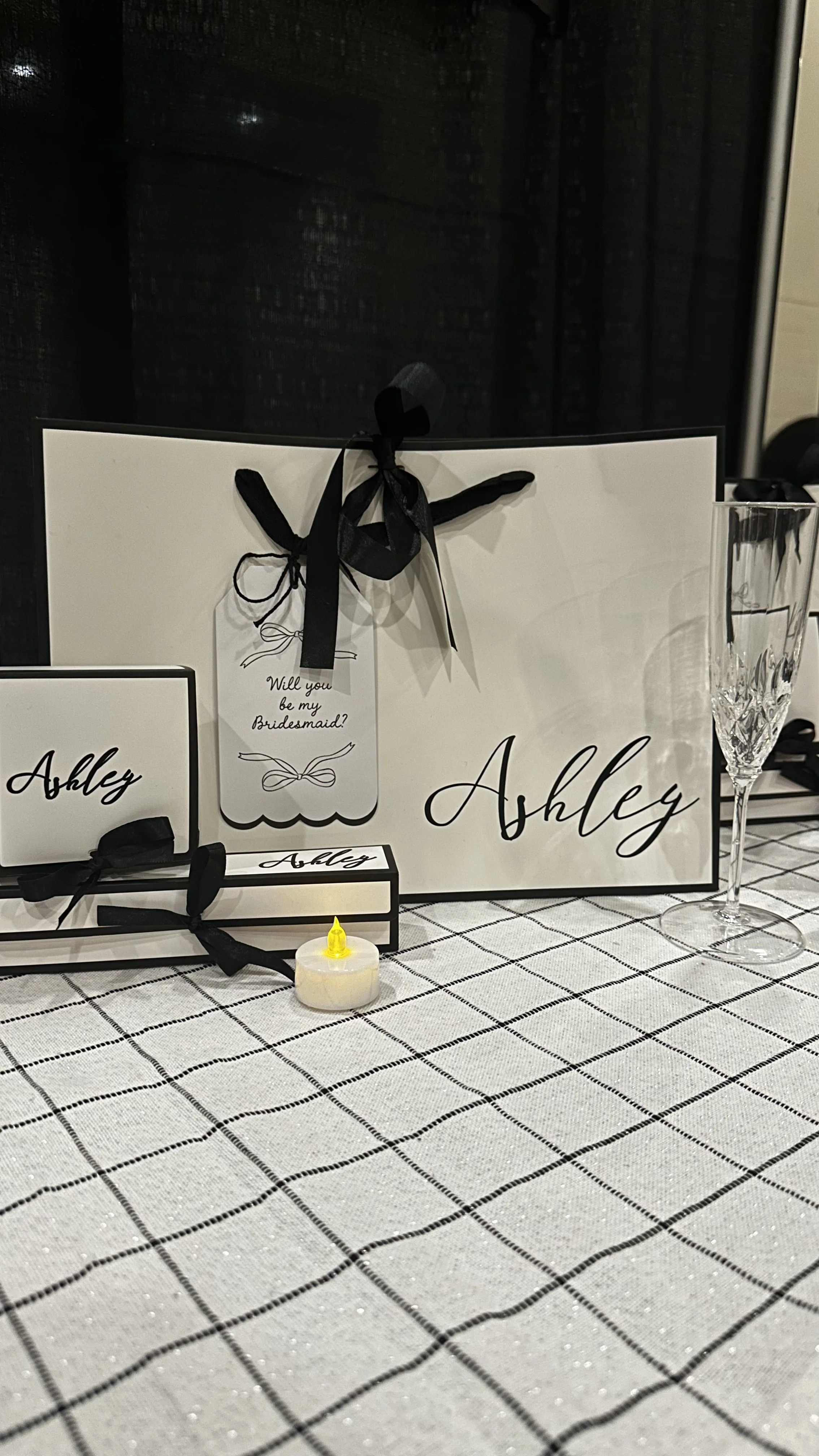 Wedding gift display with a large white sign, a smaller white box, a framed card, a small candle, and a champagne flute on a checkered tablecloth. The sign says 'Ashley' in black cursive.