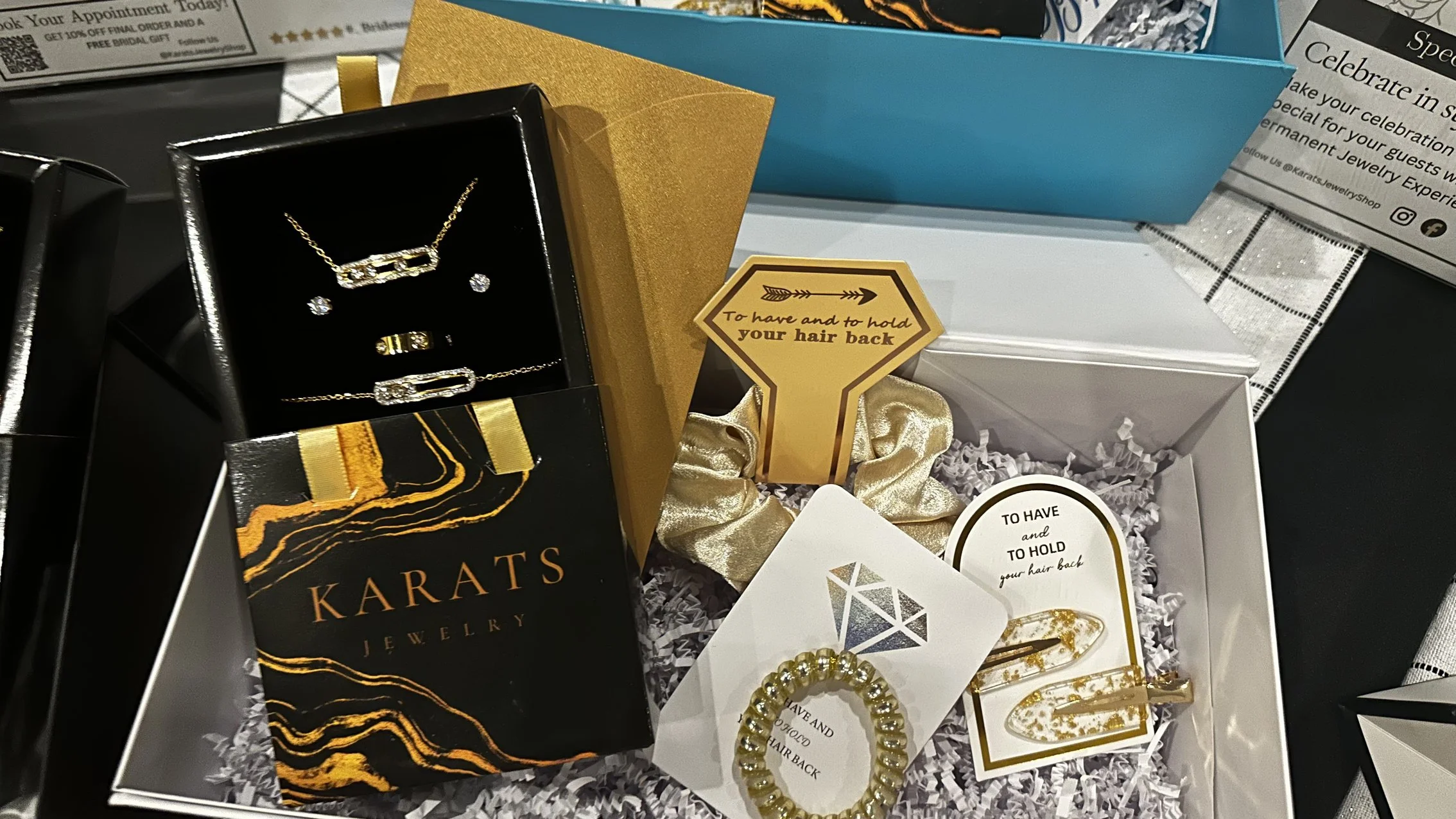 Various jewelry pieces and accessories inside gift boxes, including a necklace, earrings, a bracelet, and hair accessories, with a sign that says "To have and to hold your hair back."