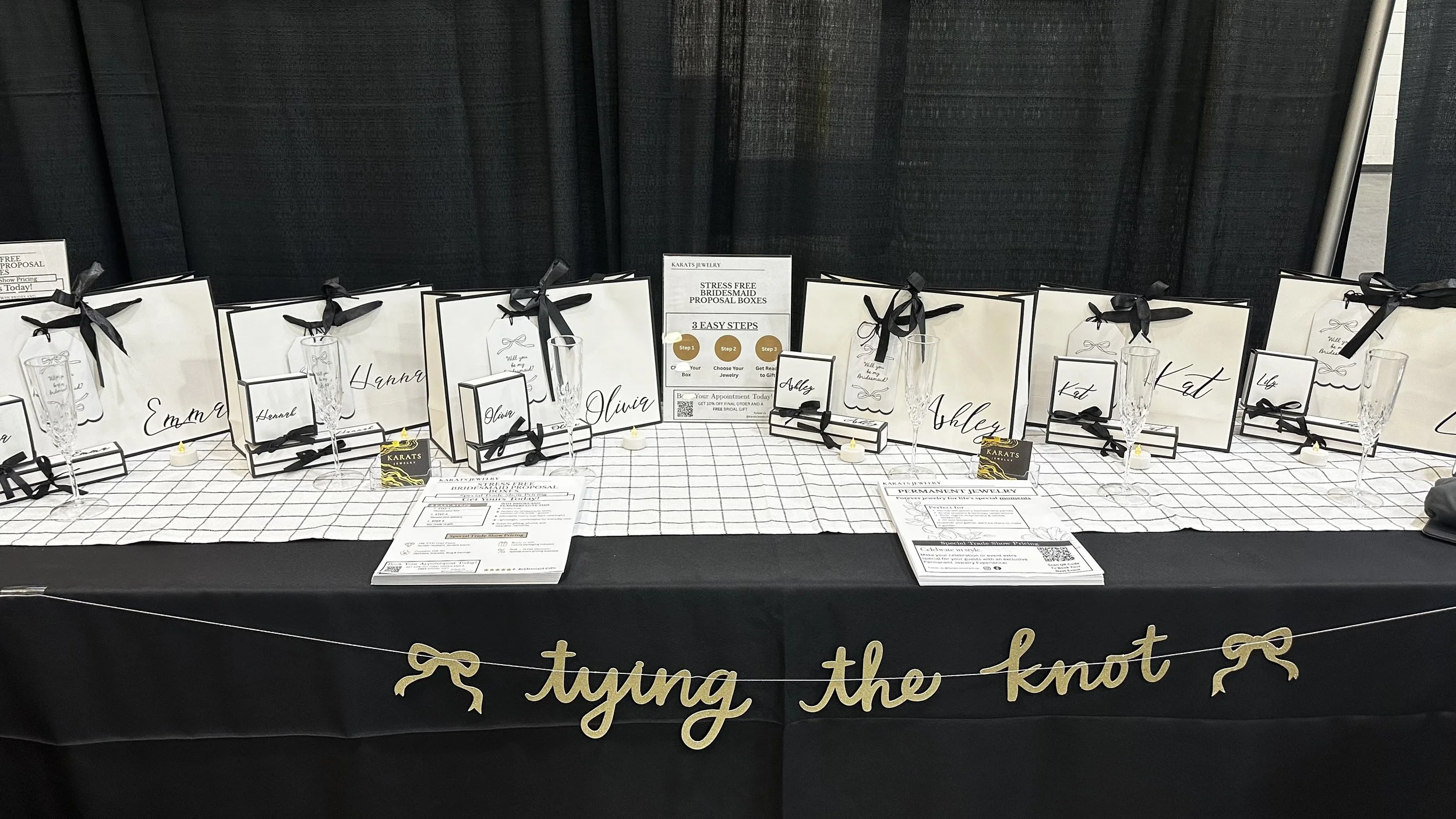 Display table showcasing jewelry and gift bags with names, promotional cards, and champagne flute glasses at a bridal event, with a black tablecloth and a gold 'tying the knot' banner.