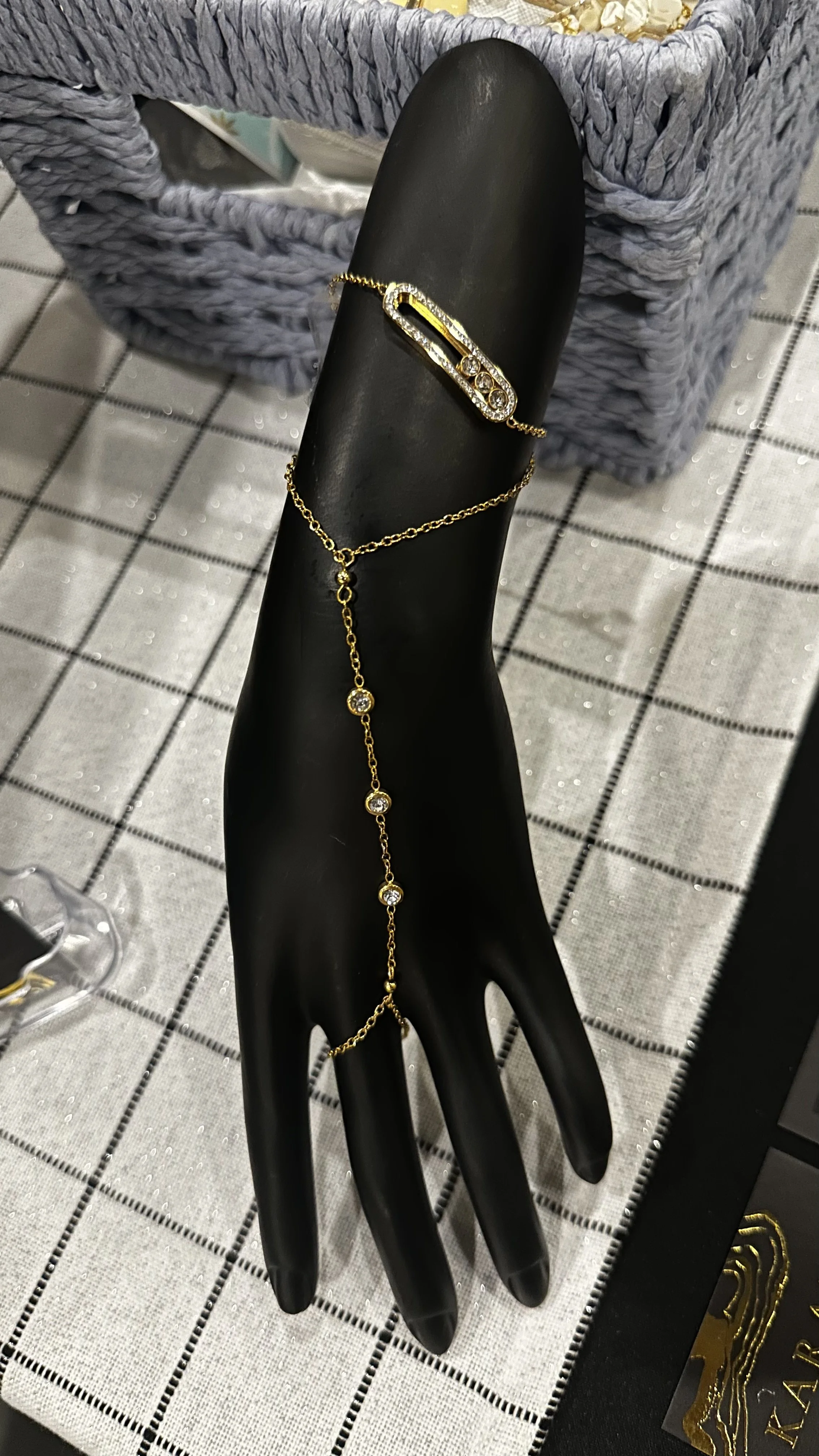 Black jewelry display hand featuring a gold bracelet with a safety pin design encrusted with rhinestones, and a gold necklace with a crime scene outline pendant and dangling rhinestones.