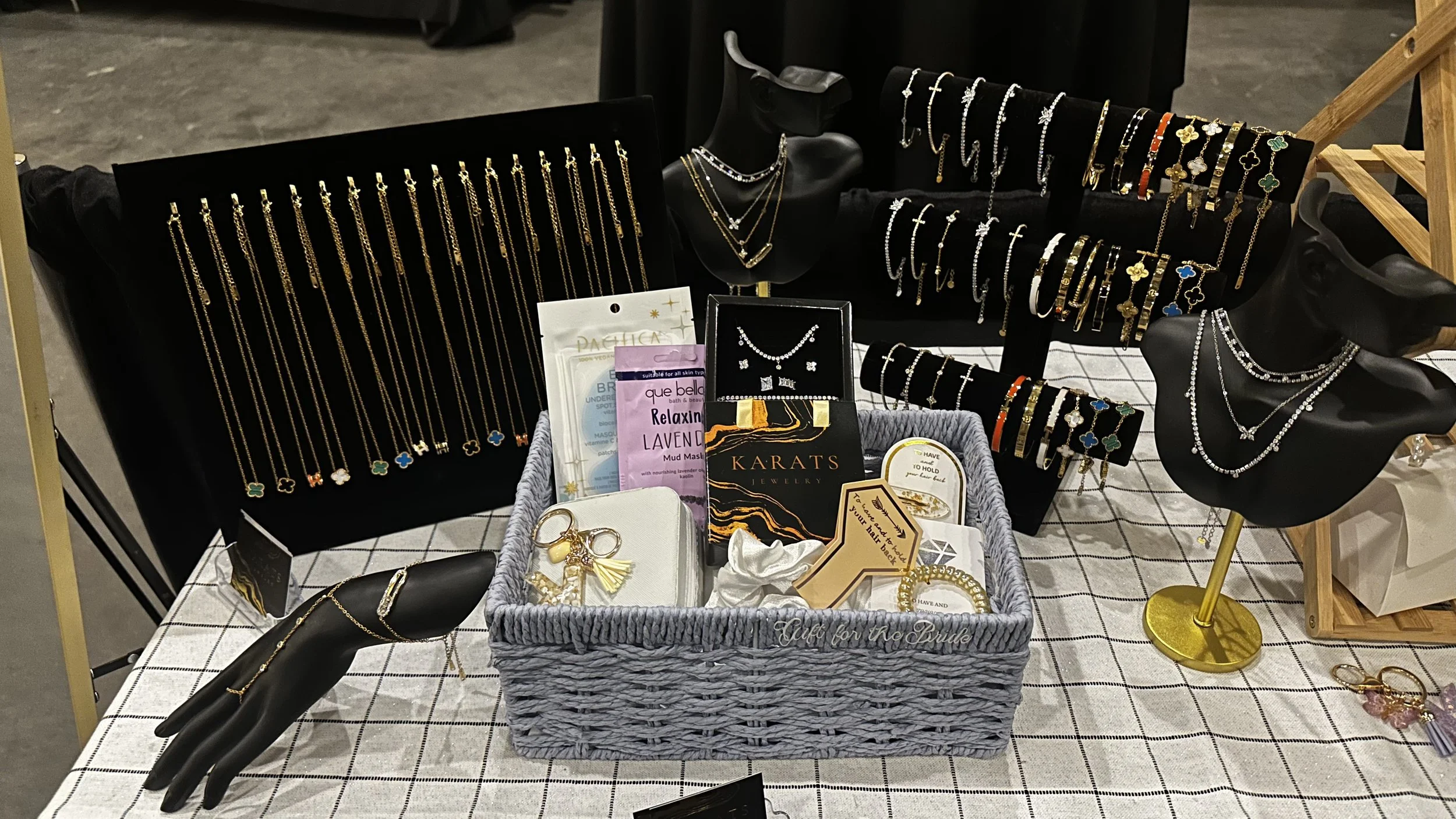 Display of various jewelry, including necklaces, bracelets, rings, and earrings on black mannequin busts, jewelry cards, and a woven basket with smaller accessories, arranged on a checkered cloth table.