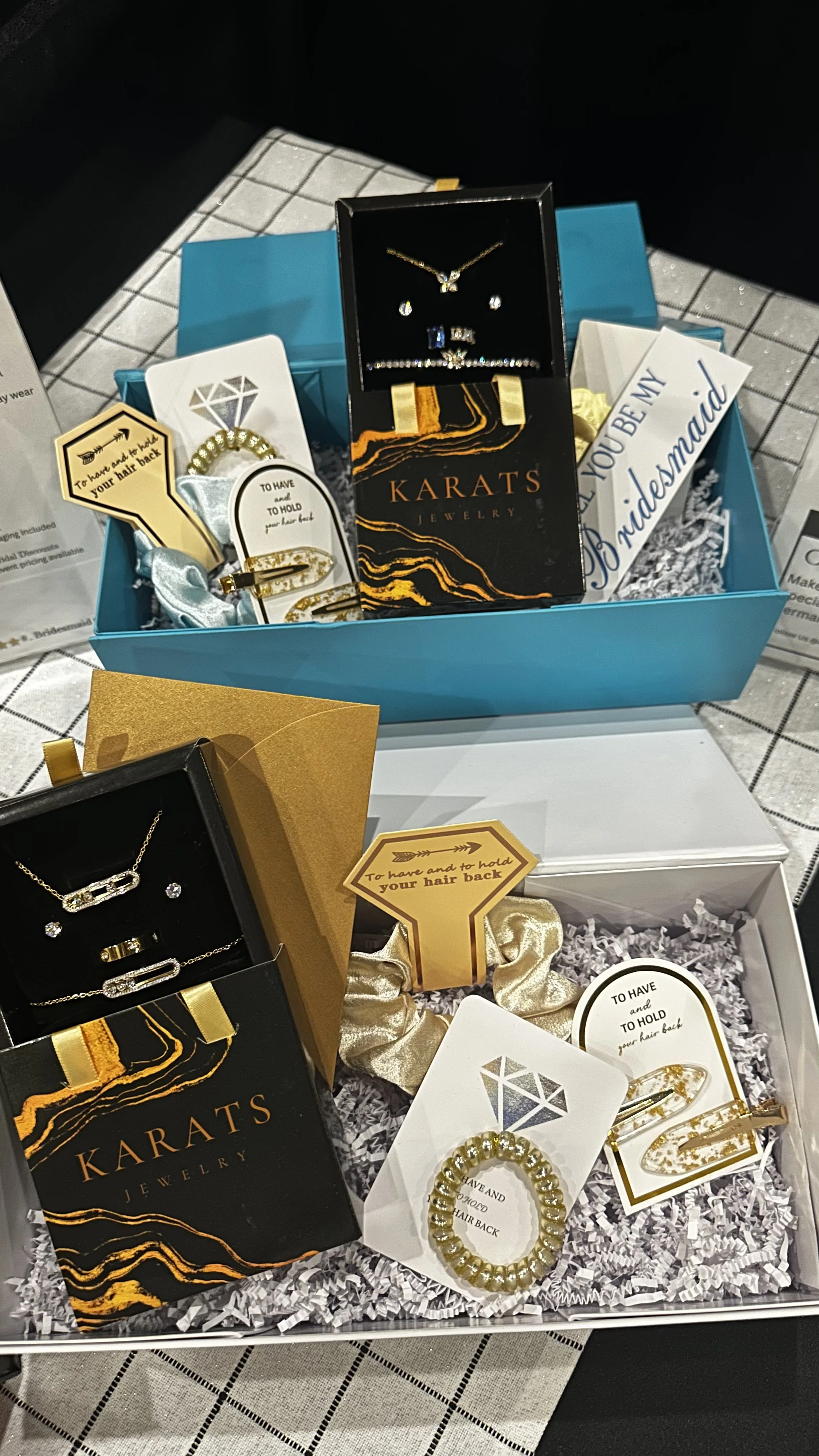 Two gift boxes with jewelry, including earrings, necklaces, bracelets, and hair accessories, on a table with a checkered tablecloth. One box is gold and the other is white, each containing jewelry and motivational or bridal-themed tags.