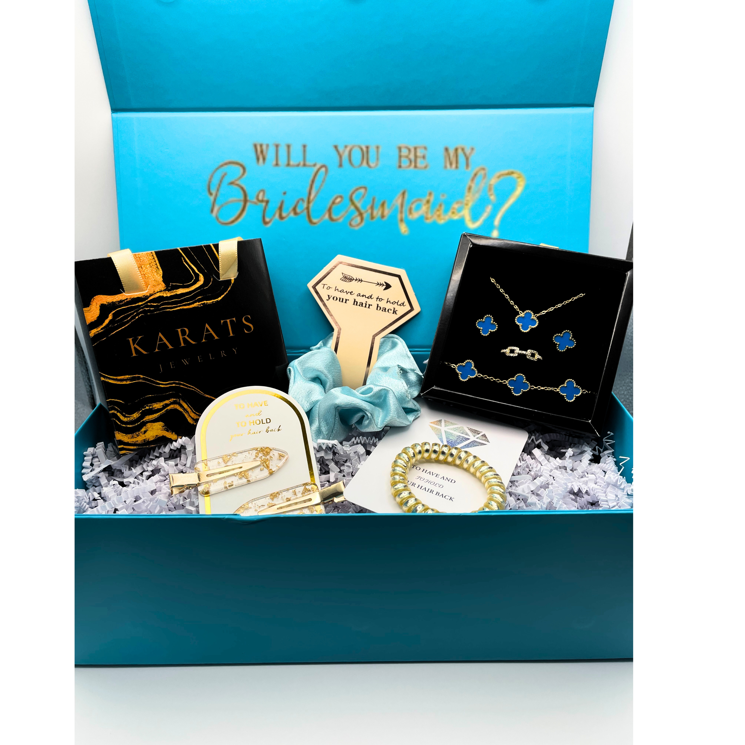 A jewelry gift box with a sign asking "Will you be my bridesmaid?" and jewelry including earrings, a necklace, a bracelet, and hair accessories with floral and gold designs.