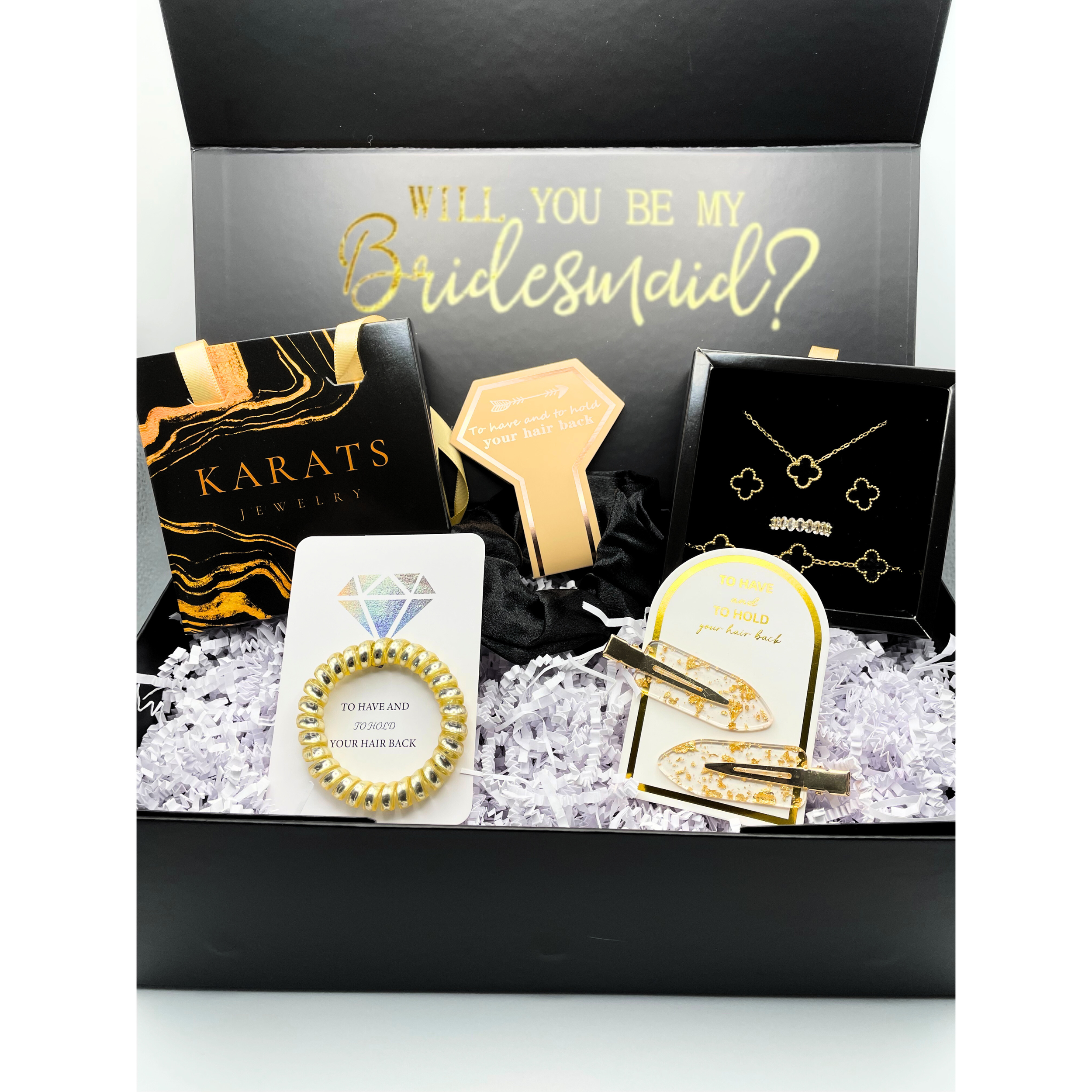 A black gift box with white paper shredded filling holds jewelry and accessories, including necklaces, hair clips, a bracelet, and a bracelet. The box features a message inside the lid reading, "Will you be my Bridesmaid?" and accessories are tagged 