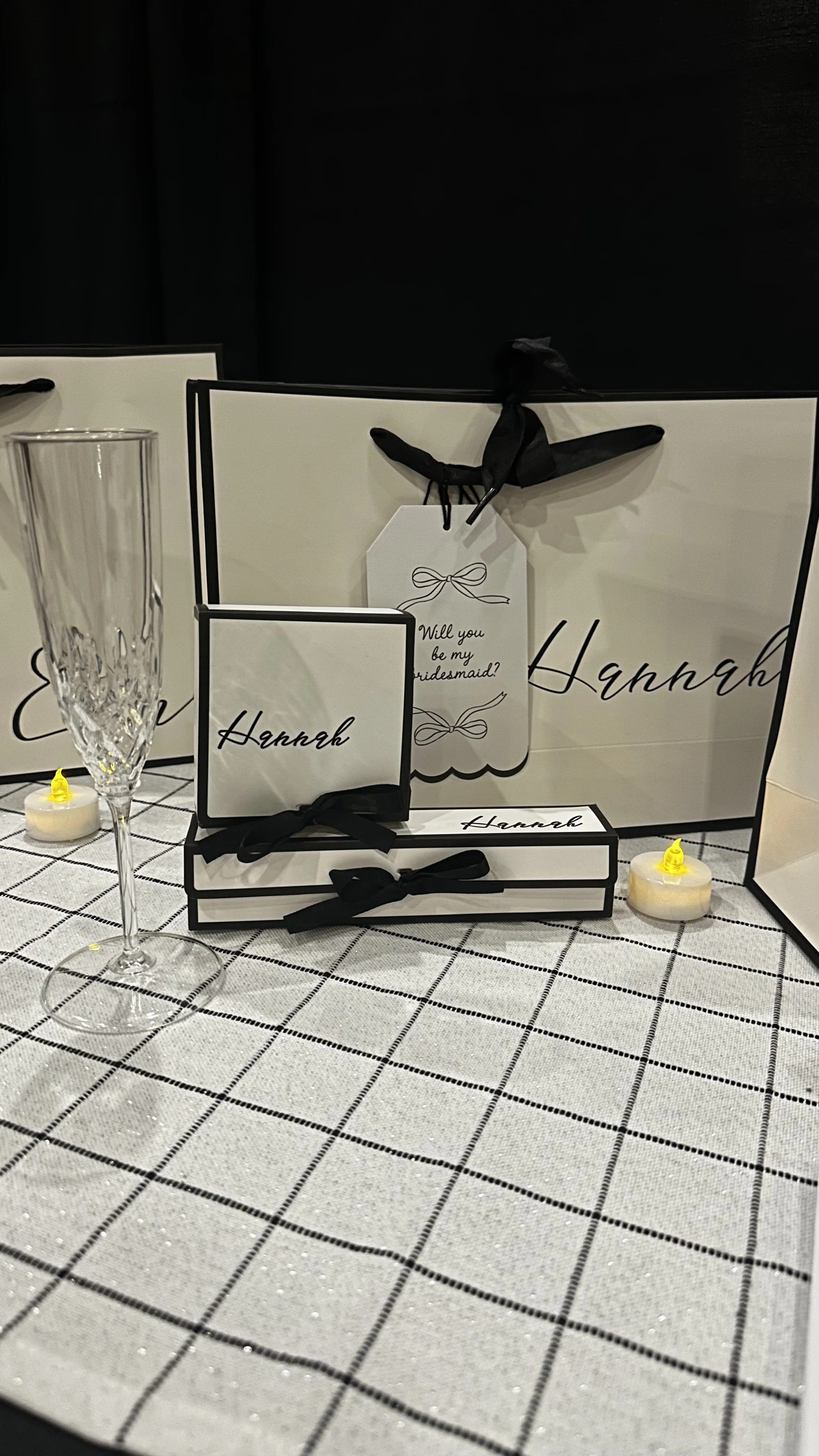 Wedding or bridal shower gift table with Hannnah-branded gift bags, boxes, and a large shopping bag, decorated with black ribbons and a tag saying "Will you be my bridesmaid?" on a checkered tablecloth, with a champagne flute and small decorative can