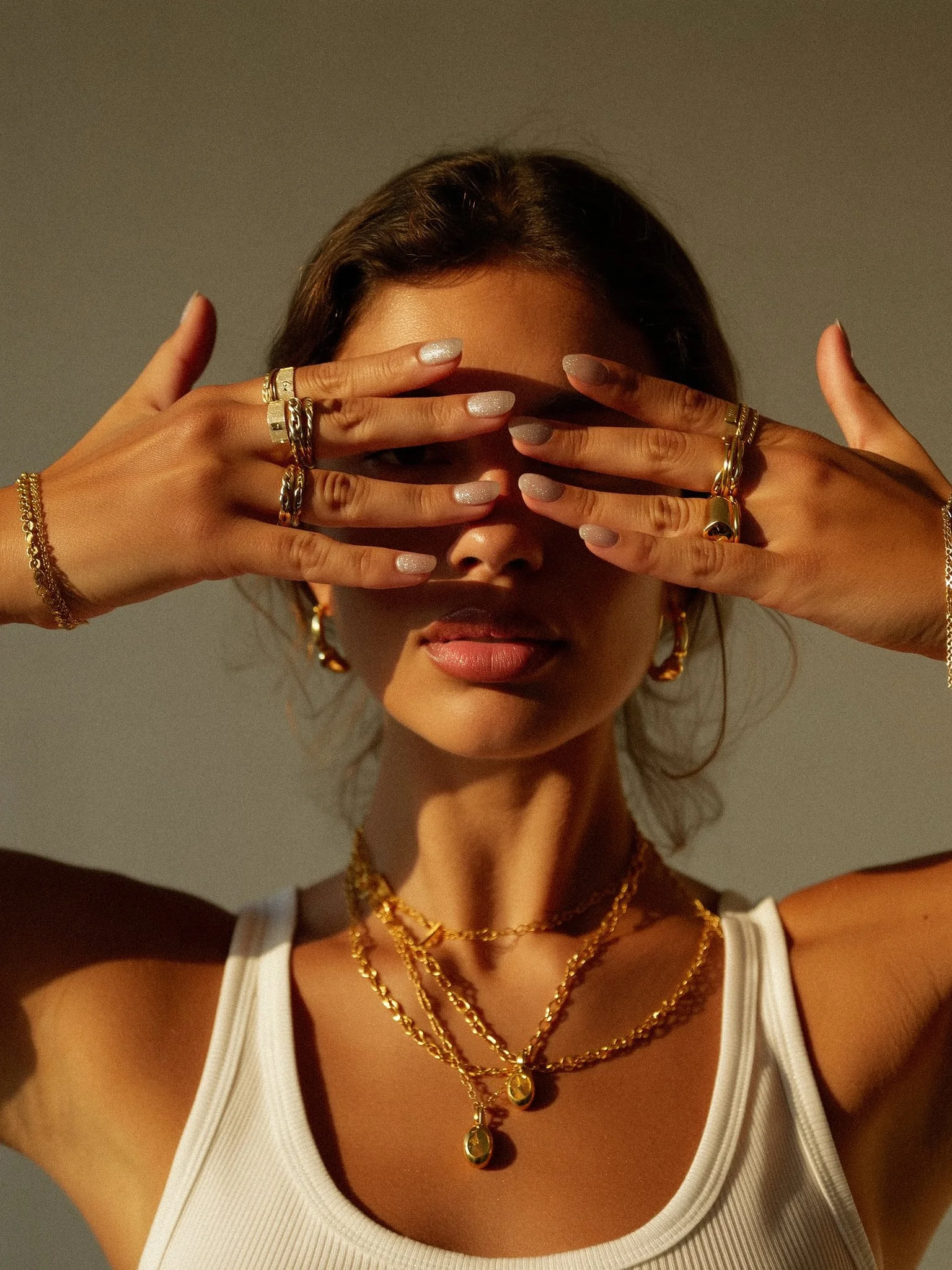 A woman with tan skin and dark hair wearing layered gold necklaces, hoop earrings, and multiple rings, holding her hands over her eyes. She has light-colored nail polish and is wearing a white tank top.