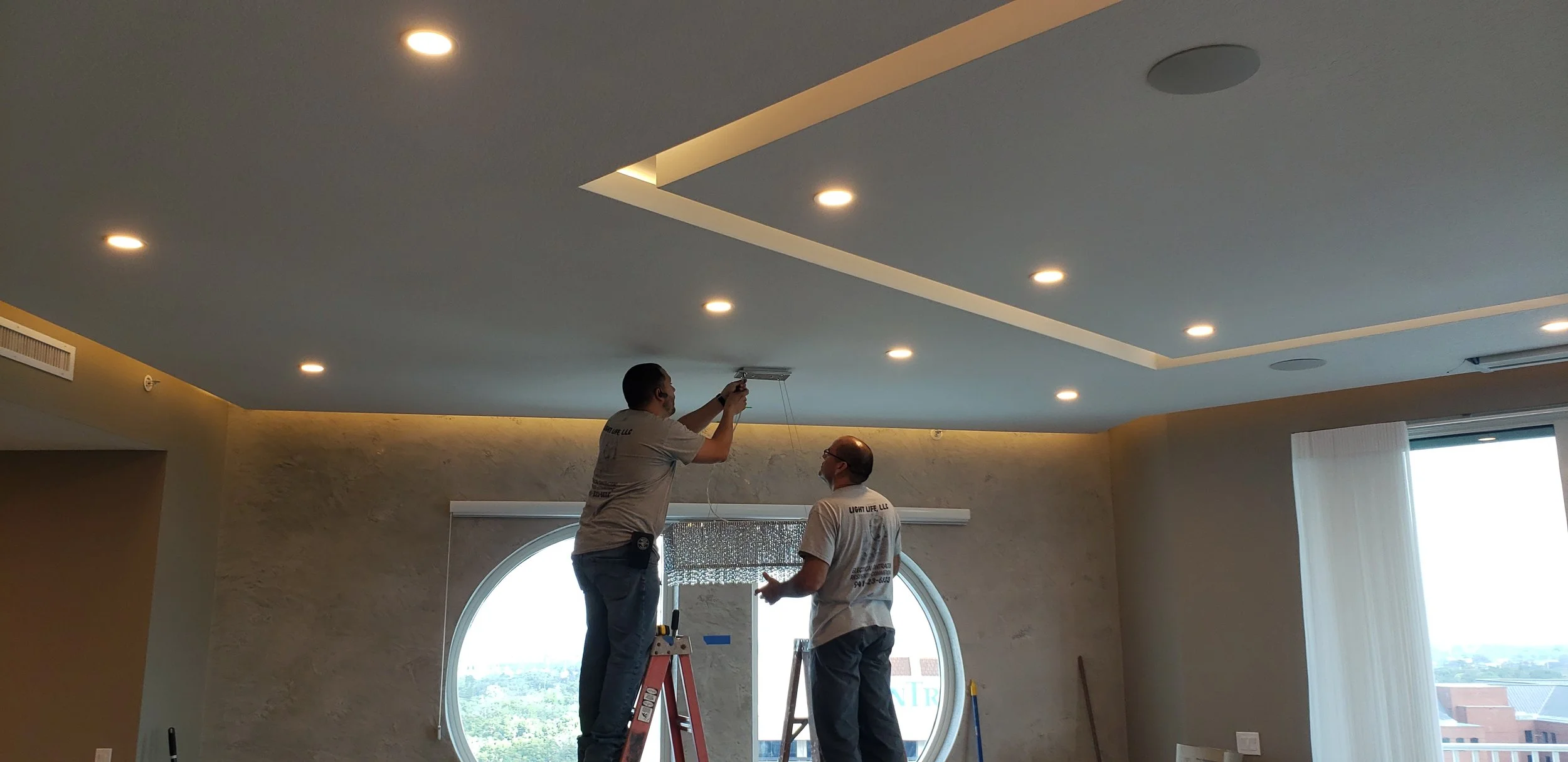 Two workers installing a ceiling light fixture in a modern room with recessed lighting and large circular window.