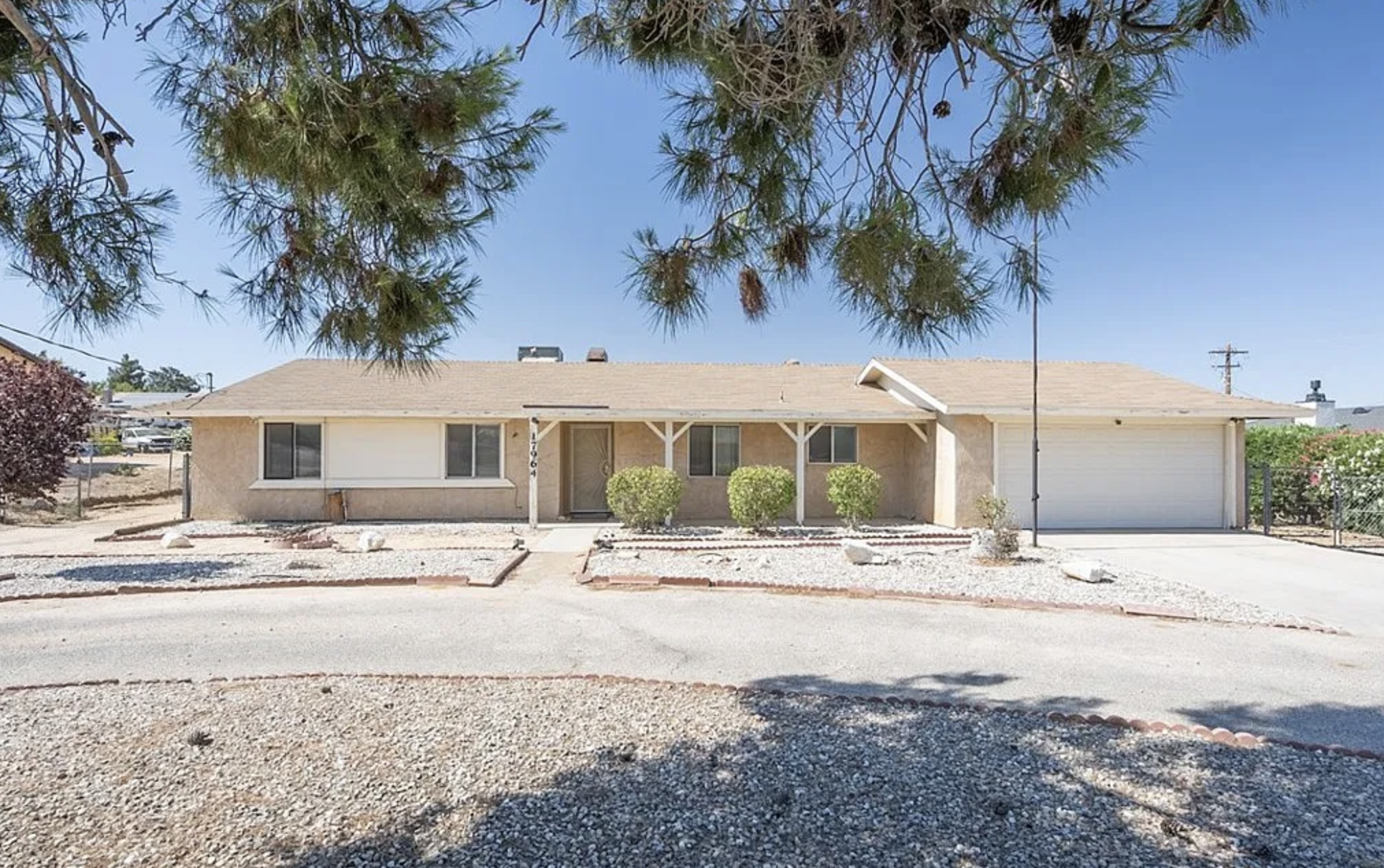 A single-story beige house with a front porch, three bushes in front, and a garage on the right, under a clear blue sky.