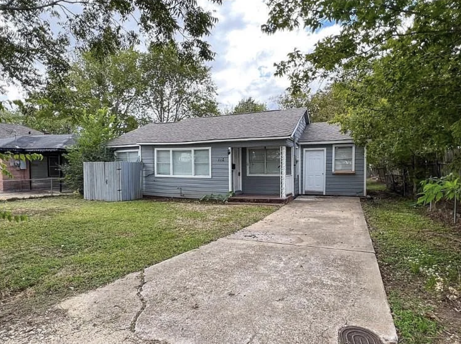A single-story house with a gray exterior and front porch, surrounded by trees and a grassy yard, with a concrete driveway leading to a side door.
