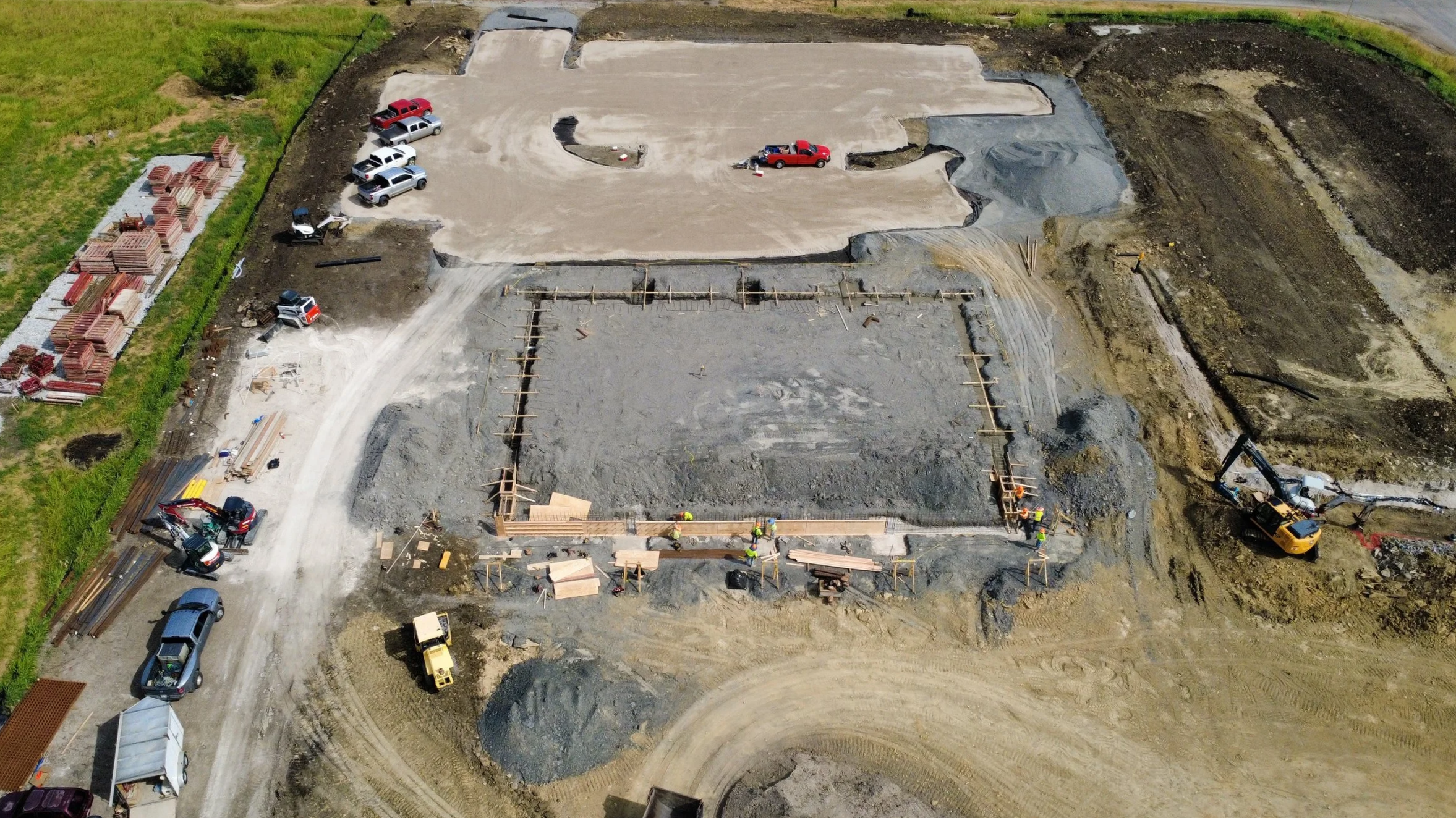 Aerial view of a construction site with foundation work in progress, several workers, construction vehicles, and parked cars surrounding the area.