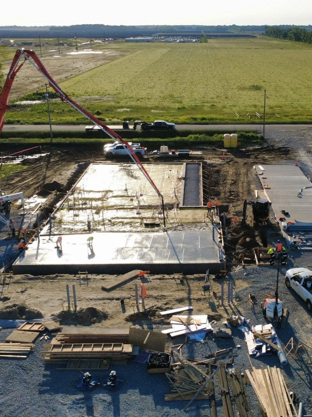 Construction site with workers pouring concrete for a building foundation, construction equipment, and materials on site. In the background, there are fields and a road with vehicles.