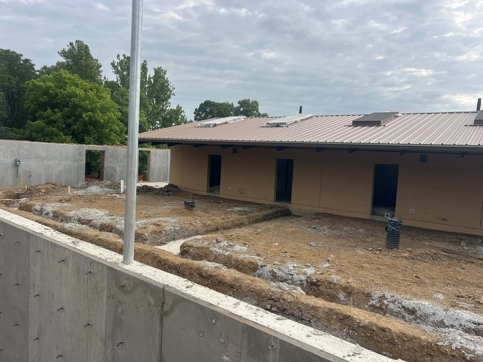 Construction site with a building that has a metal roof, surrounded by trees and a concrete wall, under cloudy sky.