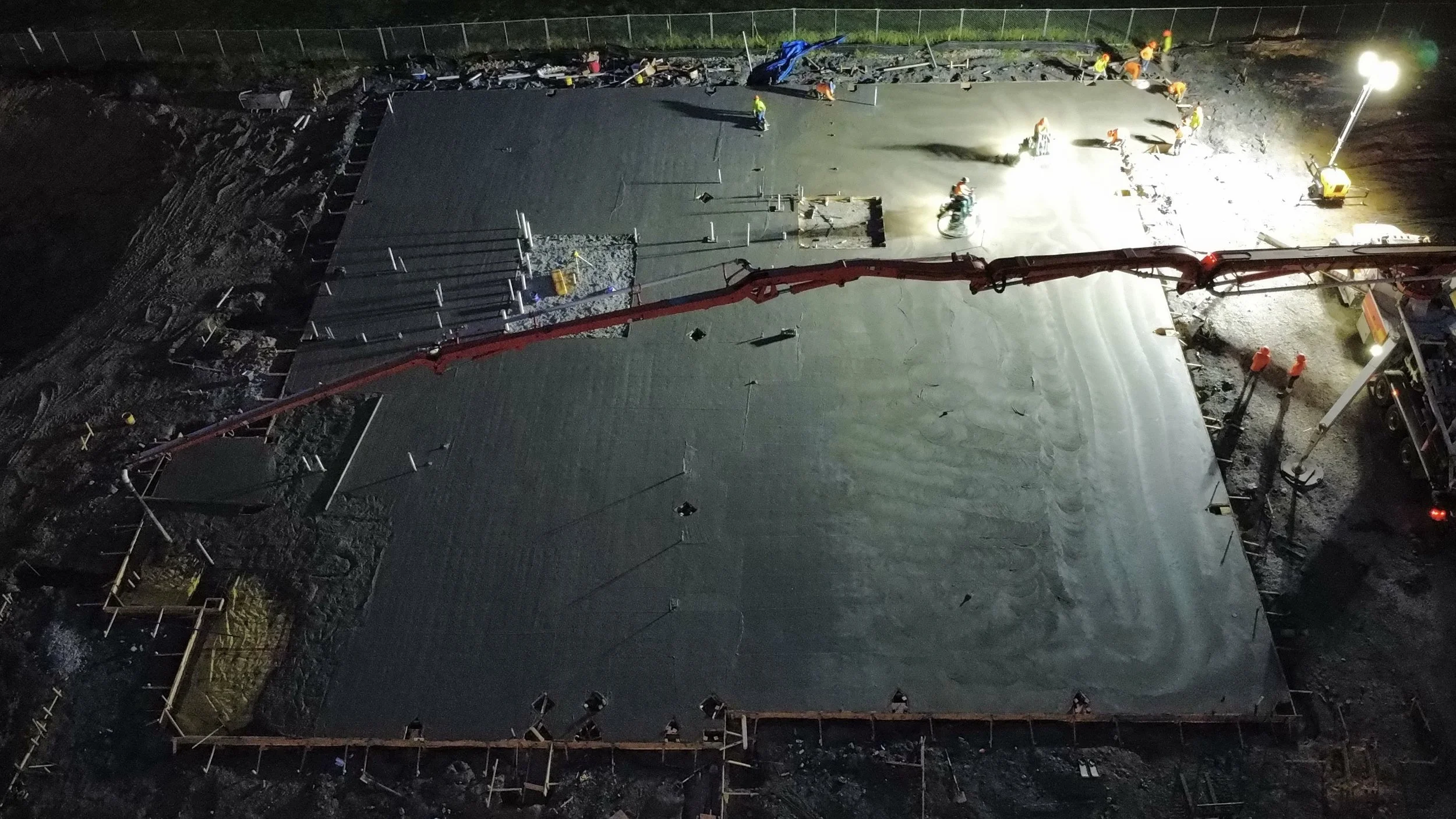 Nighttime construction site with workers pouring and smoothing concrete on a large slab, surrounded by construction equipment and safety barriers.