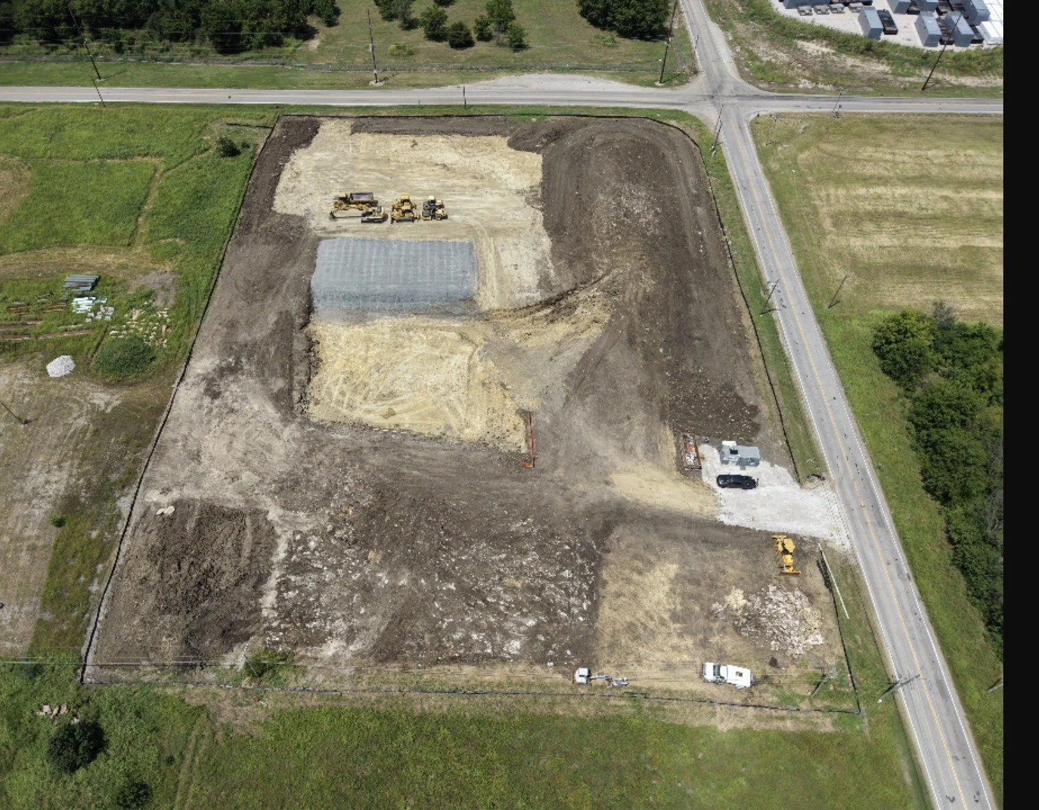 An aerial view of a construction site with excavated areas, construction vehicles, and a small greenhouse, surrounded by roads and green fields.