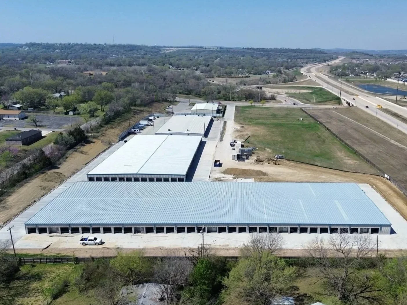 Aerial view of a large industrial warehouse complex under construction with metal roofing, surrounded by open land, roads, and some trees.
