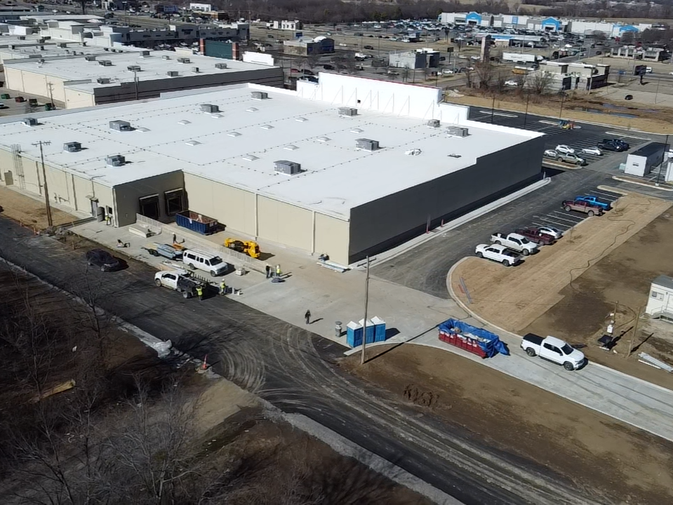 Aerial view of a large commercial building with construction activity around it, several parked cars, and construction materials nearby.