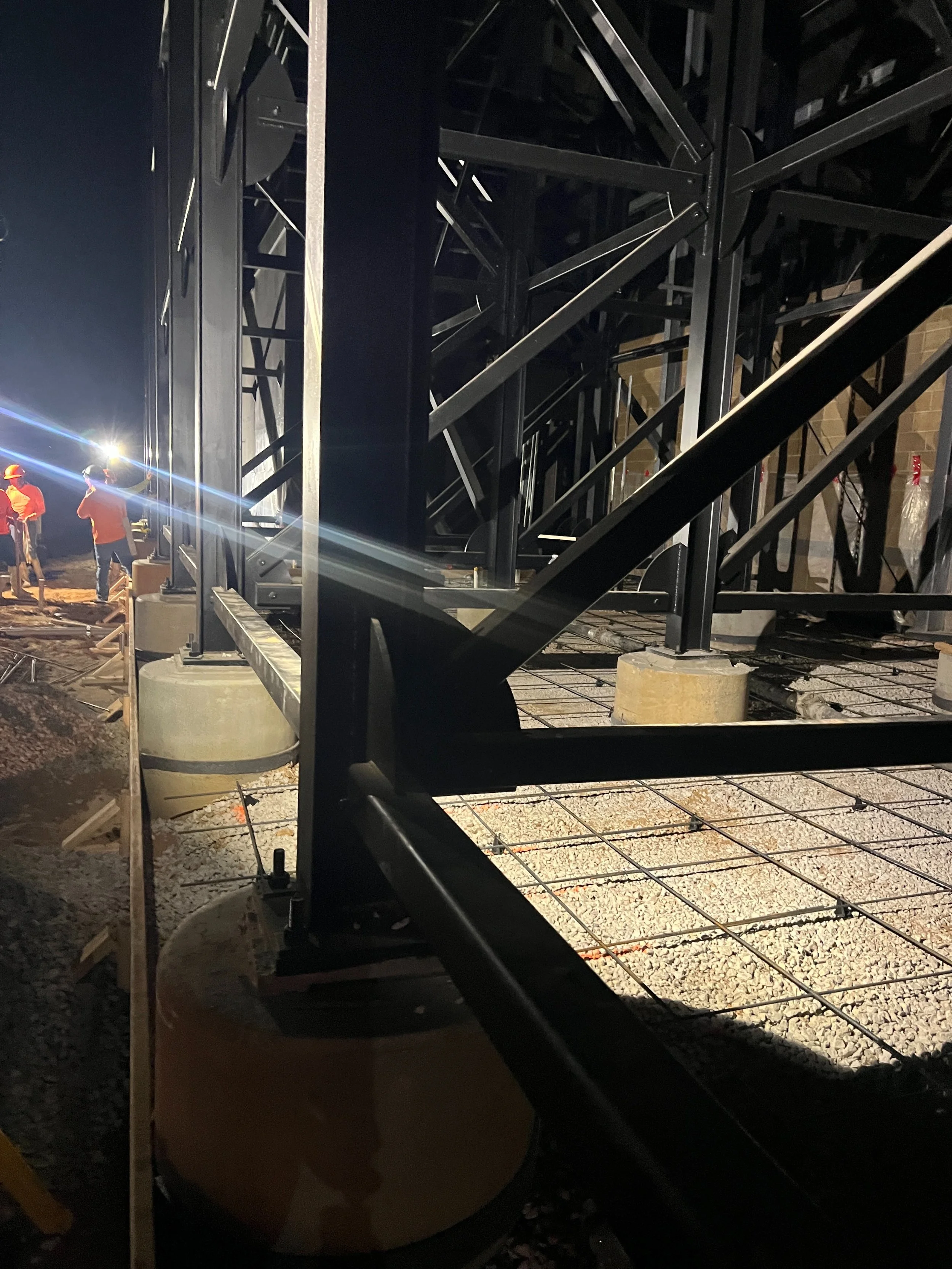 Workers at a construction site working on a steel framework structure during nighttime. The workers are wearing orange safety vests and helmets, illuminated by bright artificial lights.