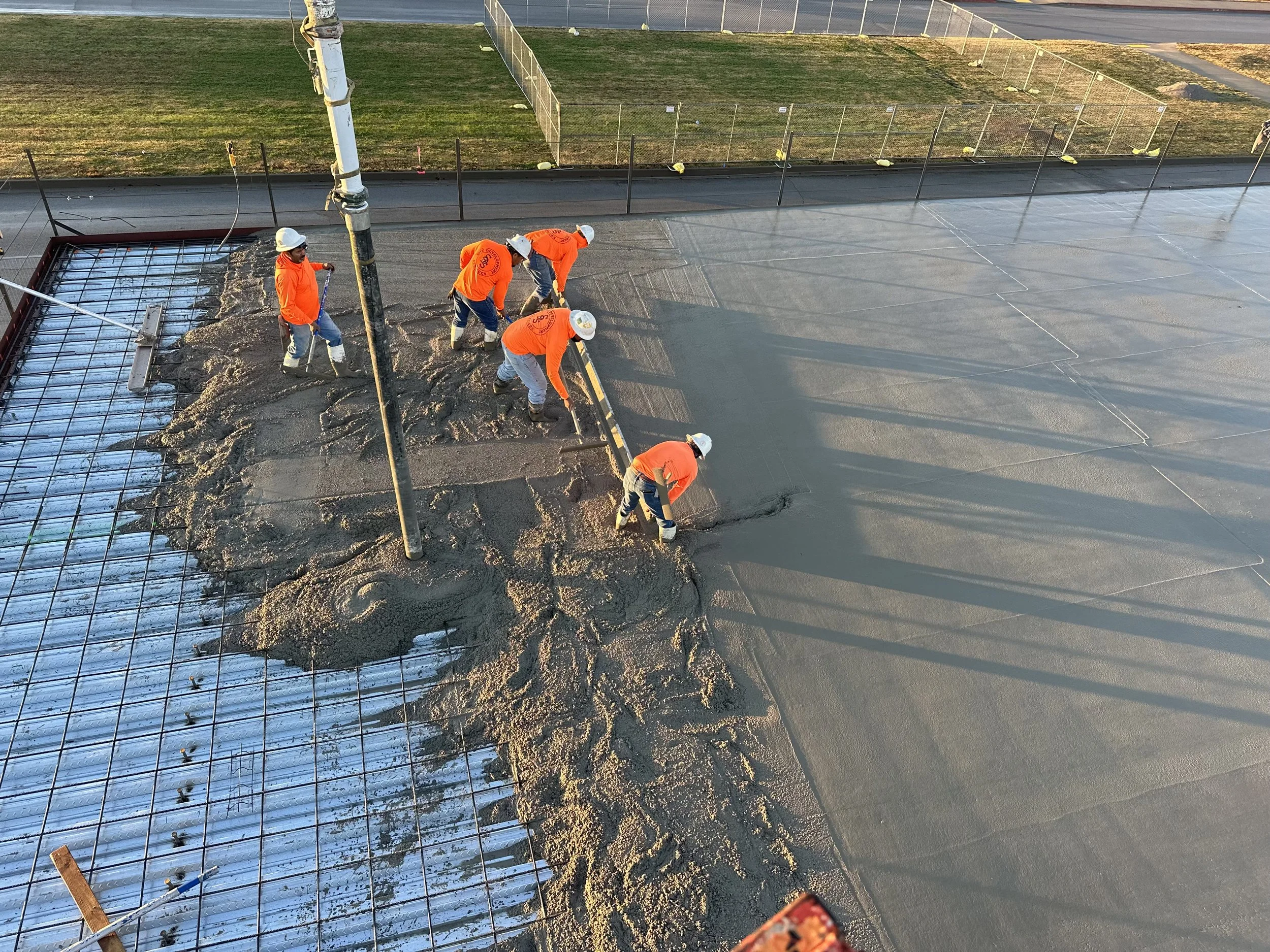Construction workers wearing orange safety jackets and white helmets pouring and leveling concrete on a building site, with a fence and grass area in the background.