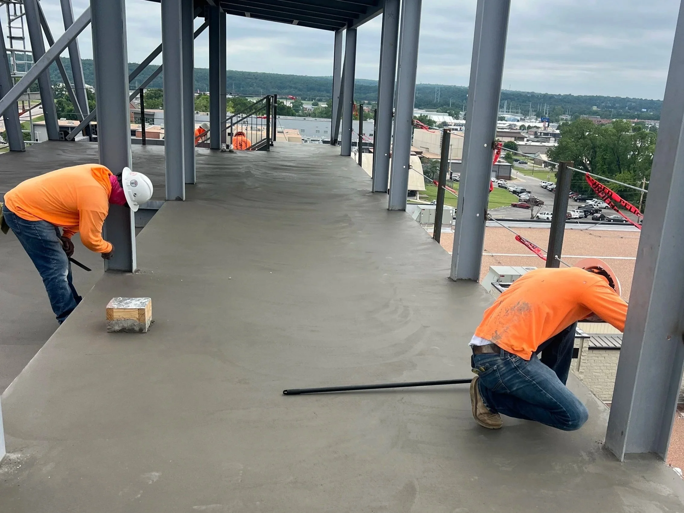 Construction workers wearing orange shirts and white safety helmets working on a building's top floor, with a cityscape and hills visible in the background.