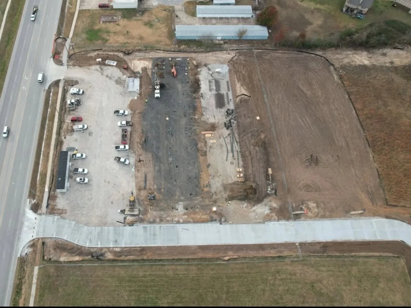 Aerial view of a construction site with dirt and gravel, construction equipment, parked vehicles, and a partially paved sidewalk, next to a road with moving cars.
