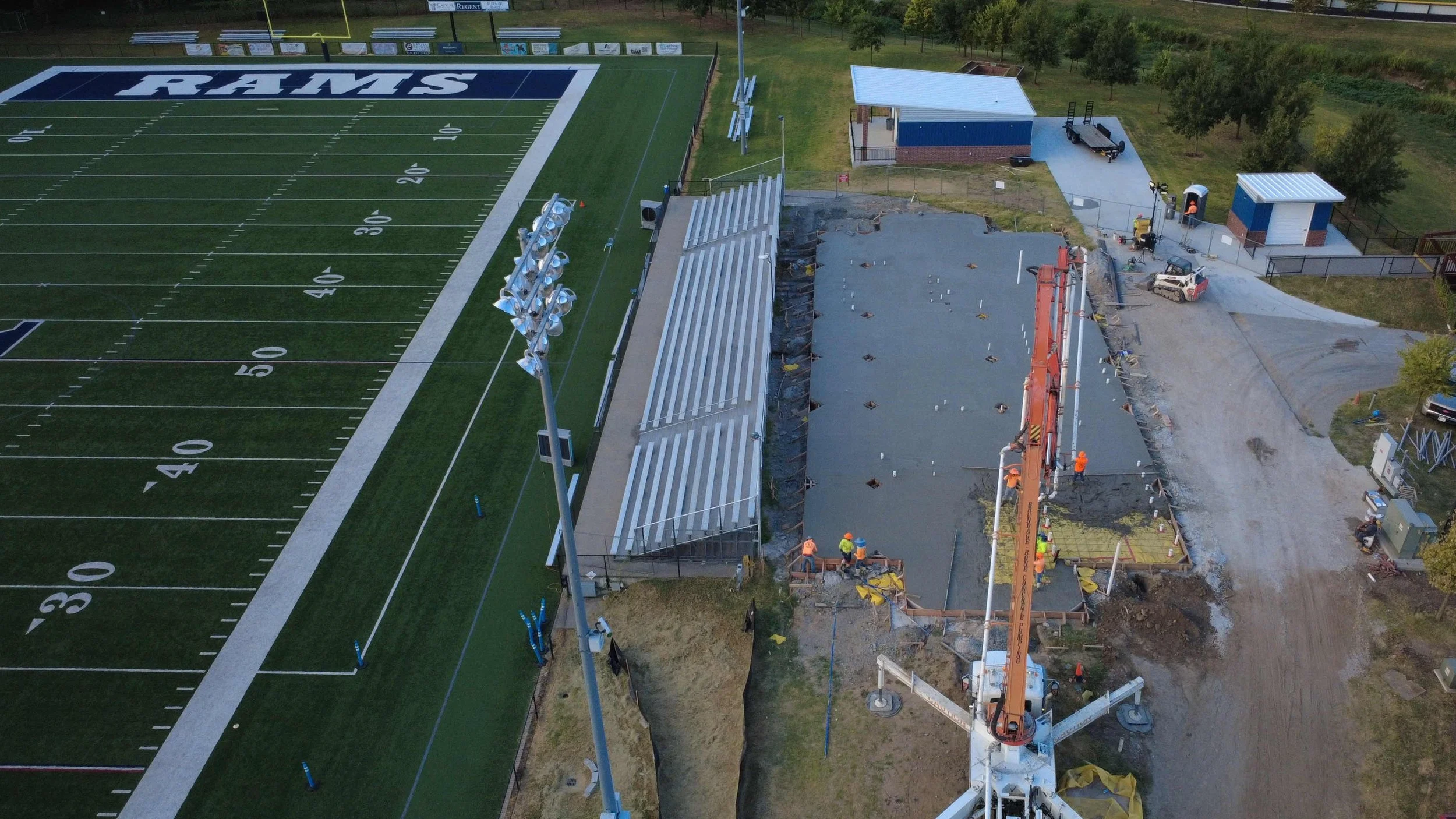 Construction workers paving a new section of stadium seating next to a football field marked with 'RAMS'.