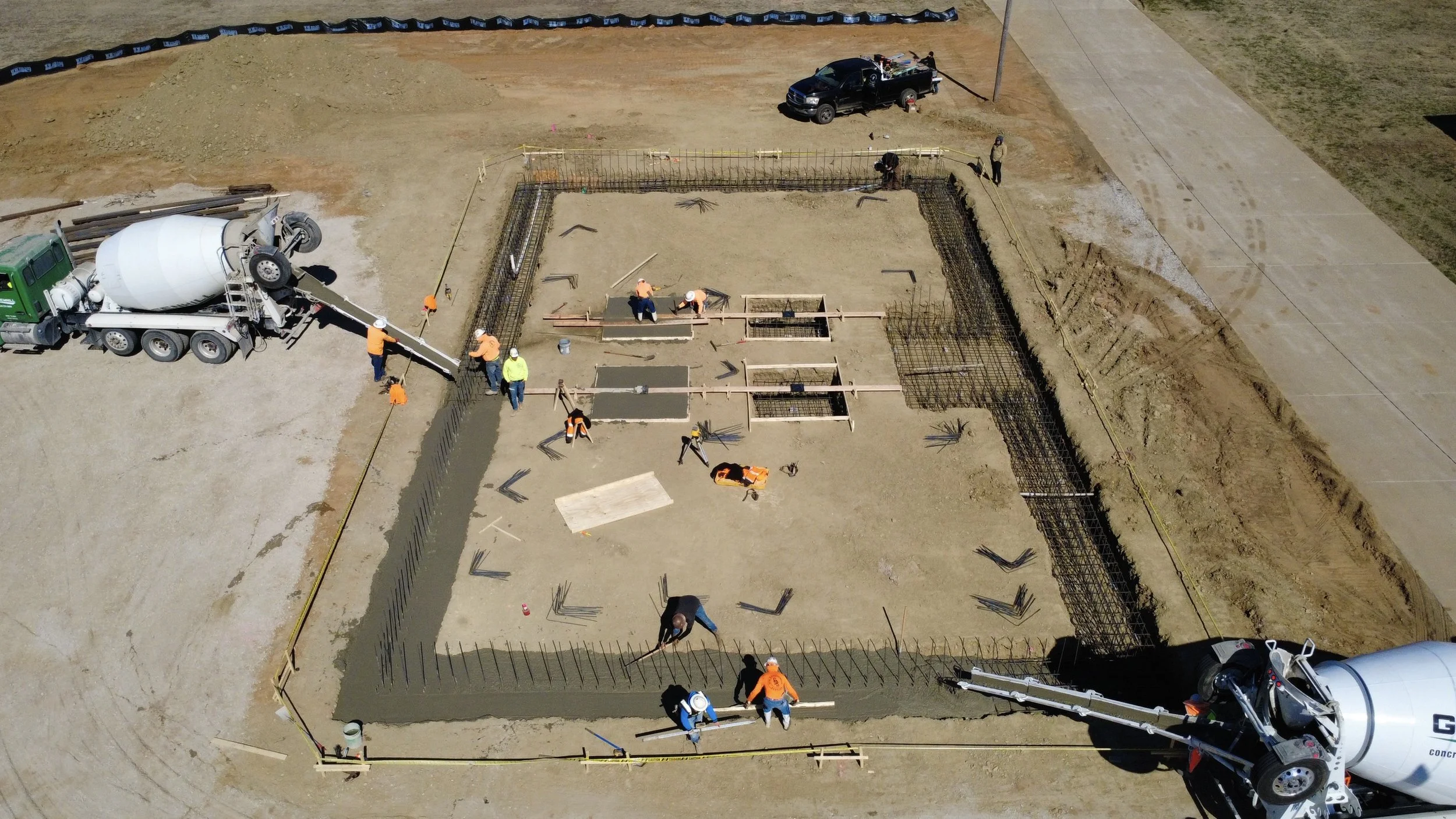 Construction workers pouring and finishing concrete foundation with rebar reinforcement in a building site, powered by a concrete mixer truck and cement truck.